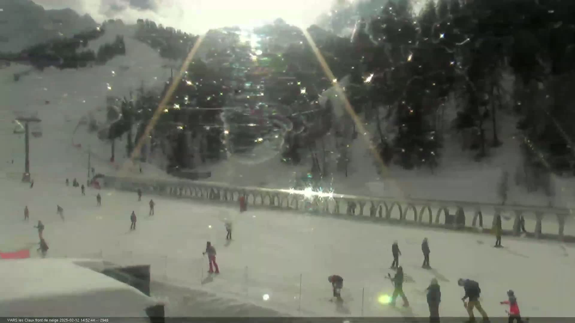 A snowy ski slope with several skiers and snowboarders visible, under a bright but somewhat overcast sky with light snow falling.