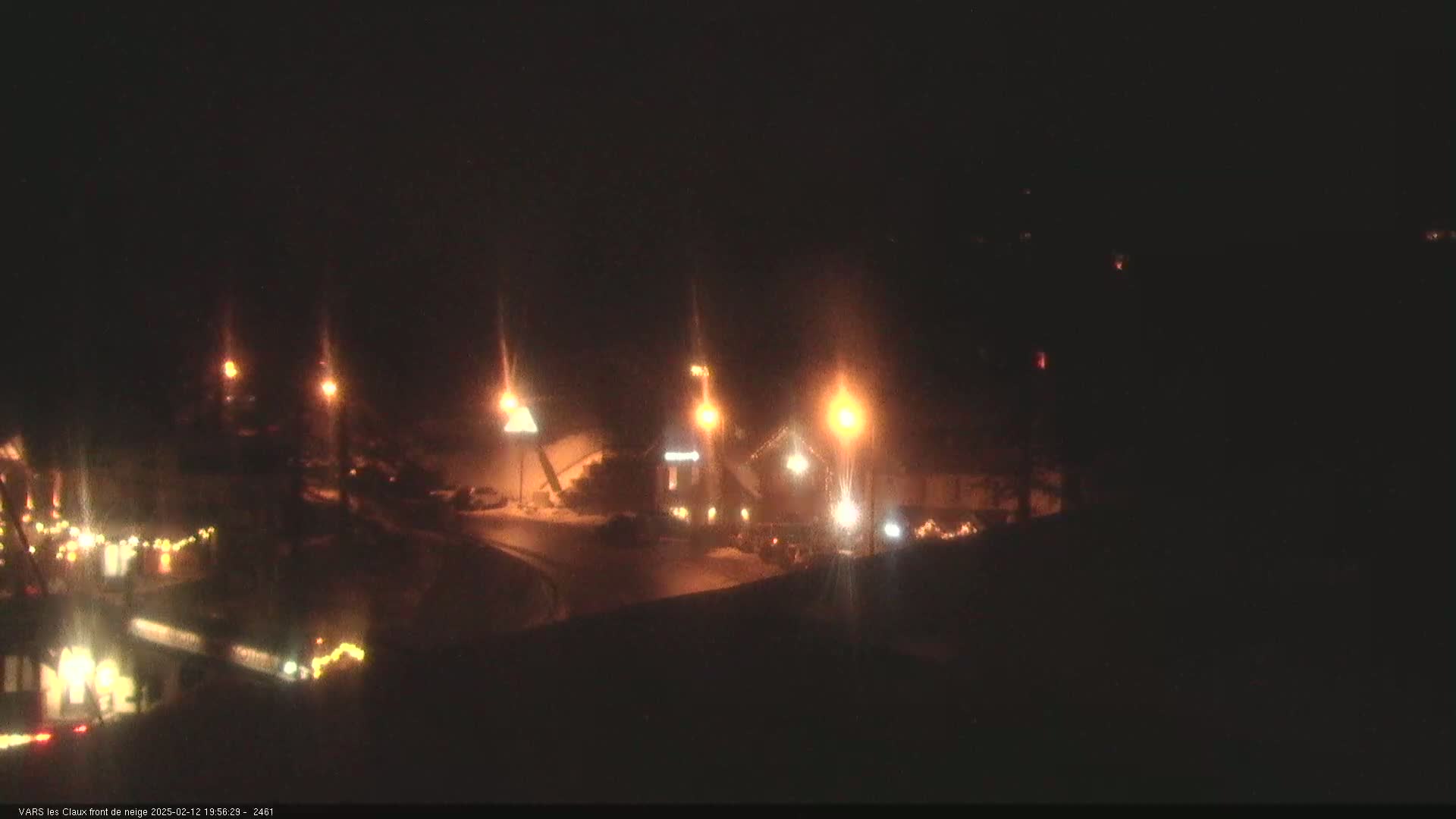 A nighttime, snowy scene shows a small town square with illuminated buildings and streetlights.