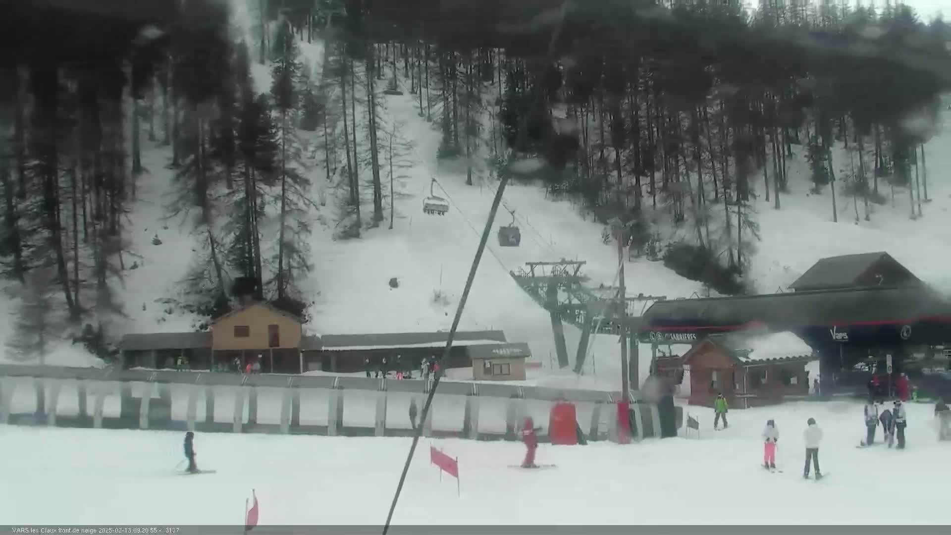 A snowy ski slope with a chairlift, several small buildings, and skiers on the piste under overcast conditions.