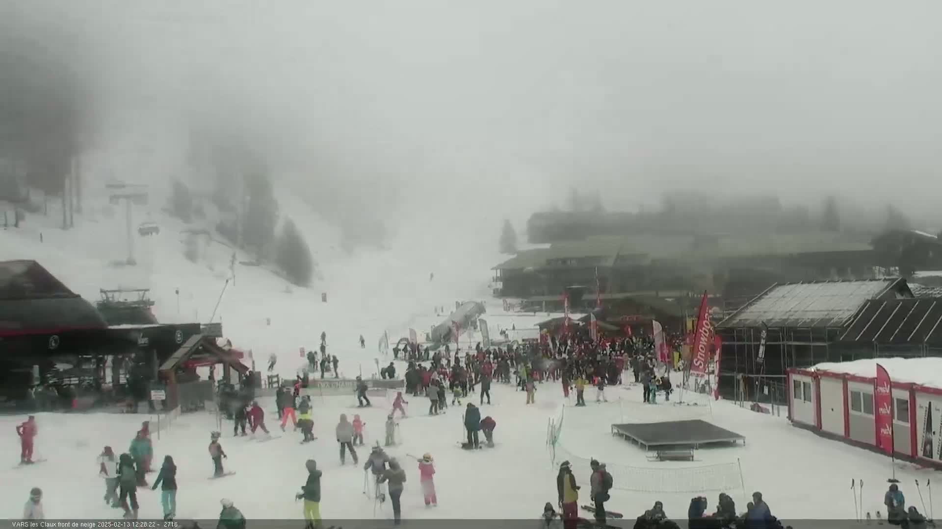 A snowy ski resort is heavily obscured by fog, with numerous skiers and snowboarders visible on the slopes and near the base area.
