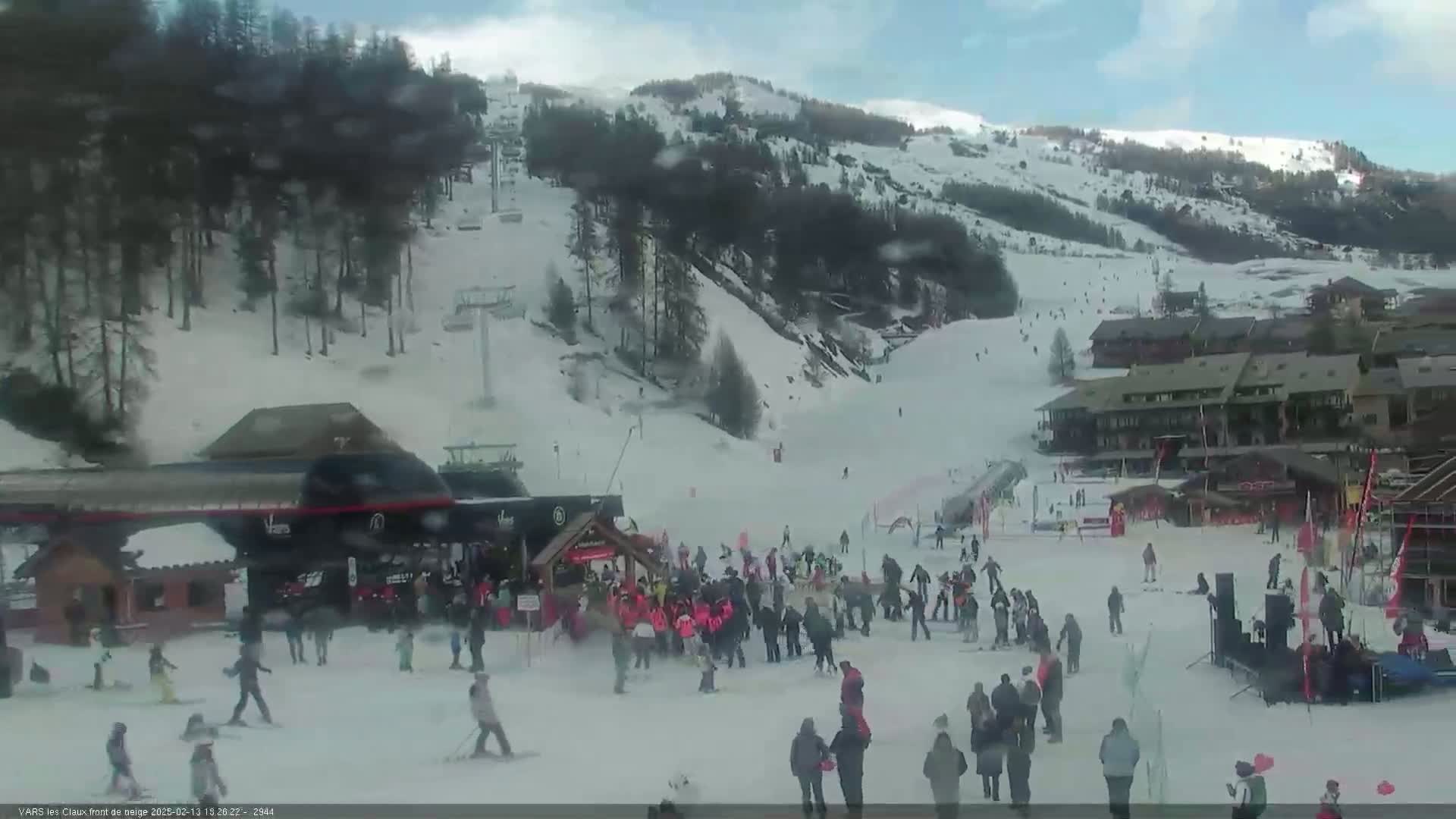 A snowy ski resort with a crowd of people gathered near a building at the base of a ski slope on a somewhat cloudy day.