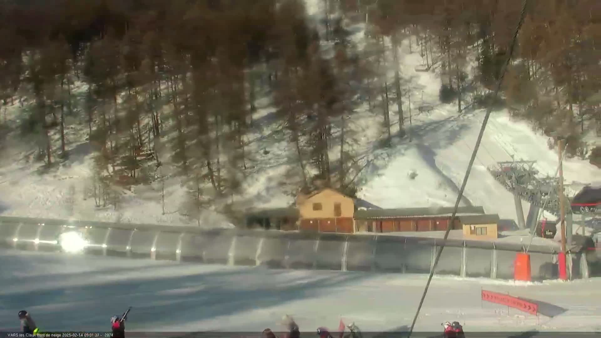A sunny day at a snow-covered ski resort shows a ski lift, a building, and several skiers at the bottom of a slope.