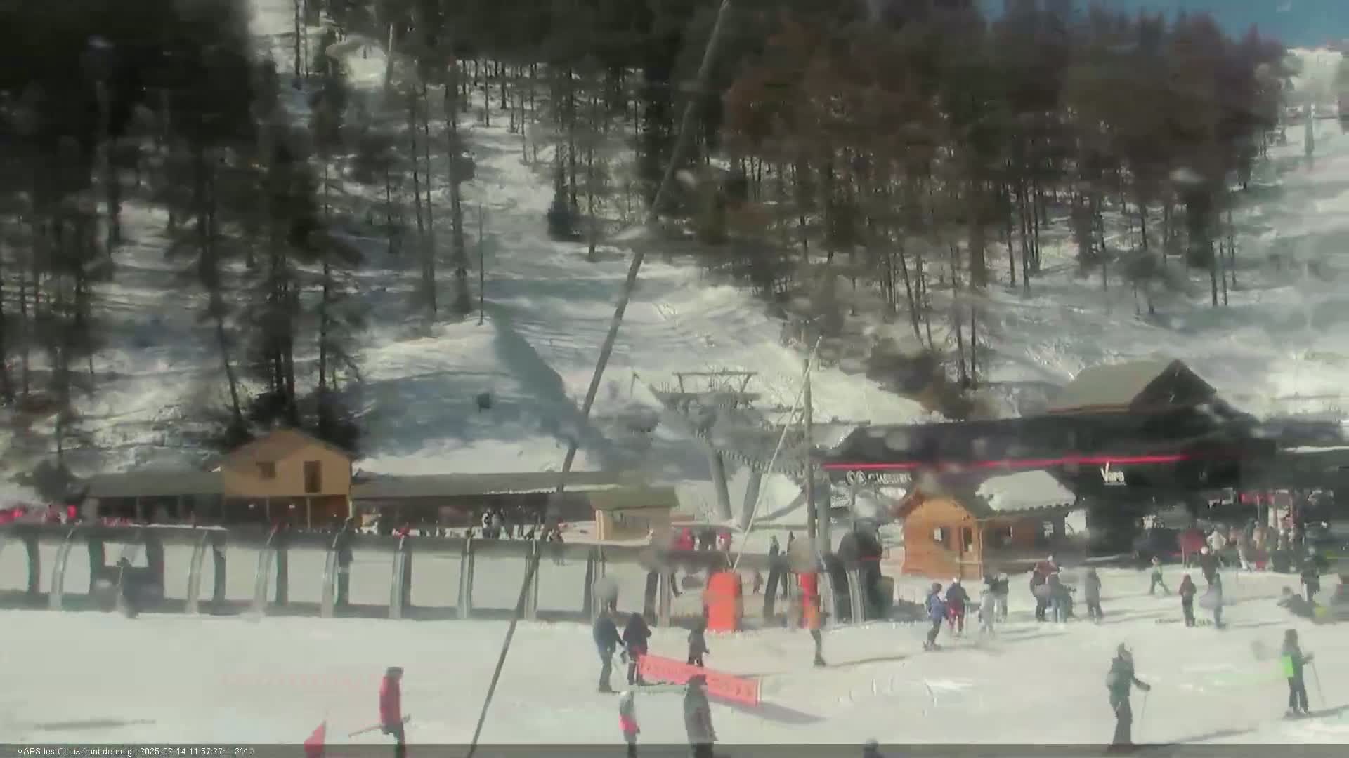 A sunny day at a snow-covered ski resort shows a chairlift, several buildings, and skiers and snowboarders on the slopes and near the base.