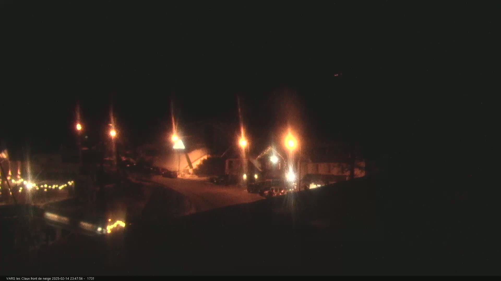 At night, a snow-covered area is illuminated by several streetlights, revealing buildings and vehicles.