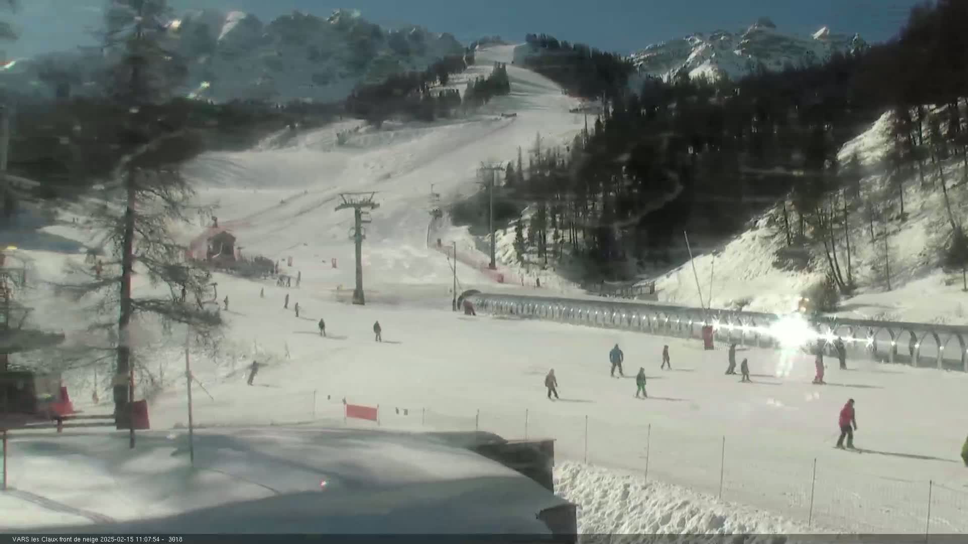 A sunny day at a snow-covered ski resort shows several skiers and snowboarders on the slopes, with mountains in the background and ski lifts visible.