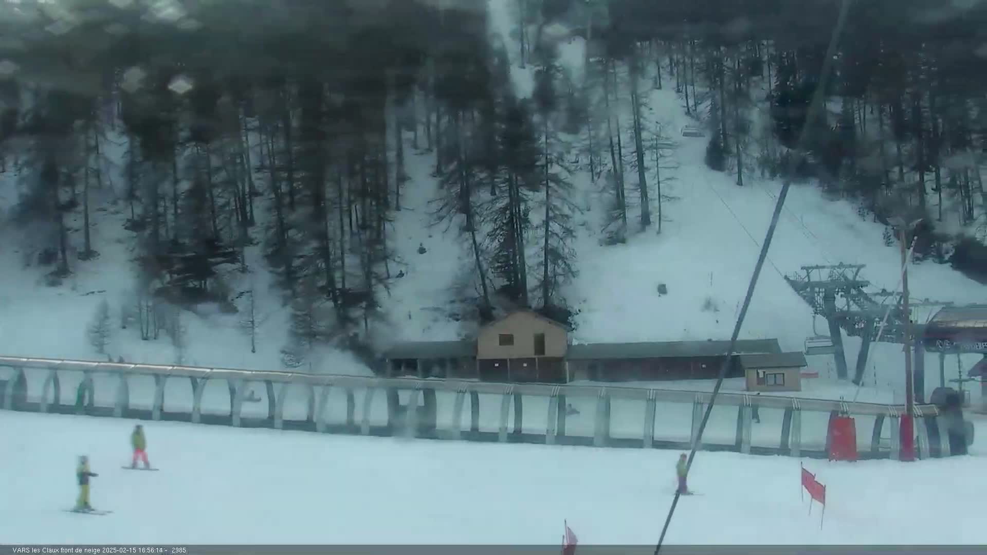 A snowy ski slope with a chairlift and a few skiers, partially obscured by blowing snow, is situated below a forested hillside.