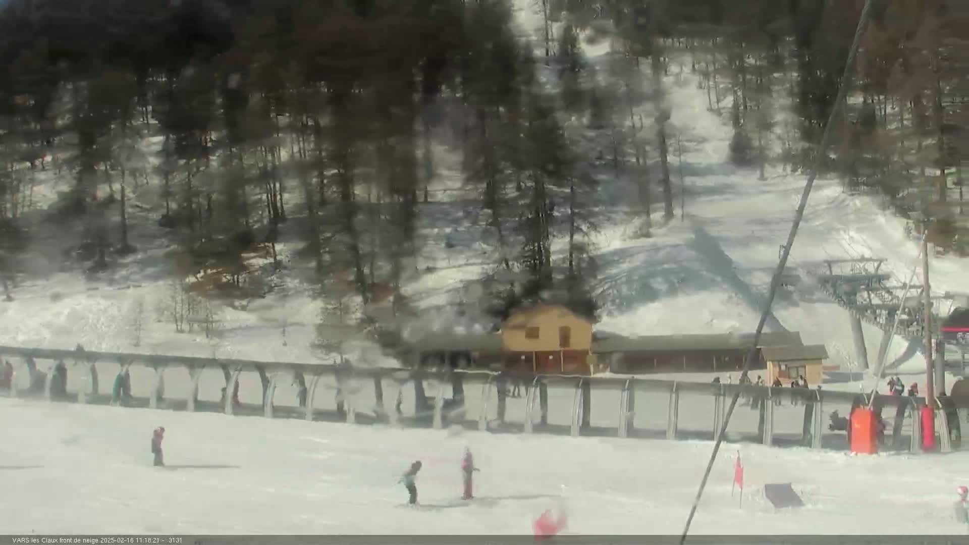 A snowy ski slope with a few skiers and snowboarders, a small building, and a chairlift in the background on a bright, somewhat hazy day.