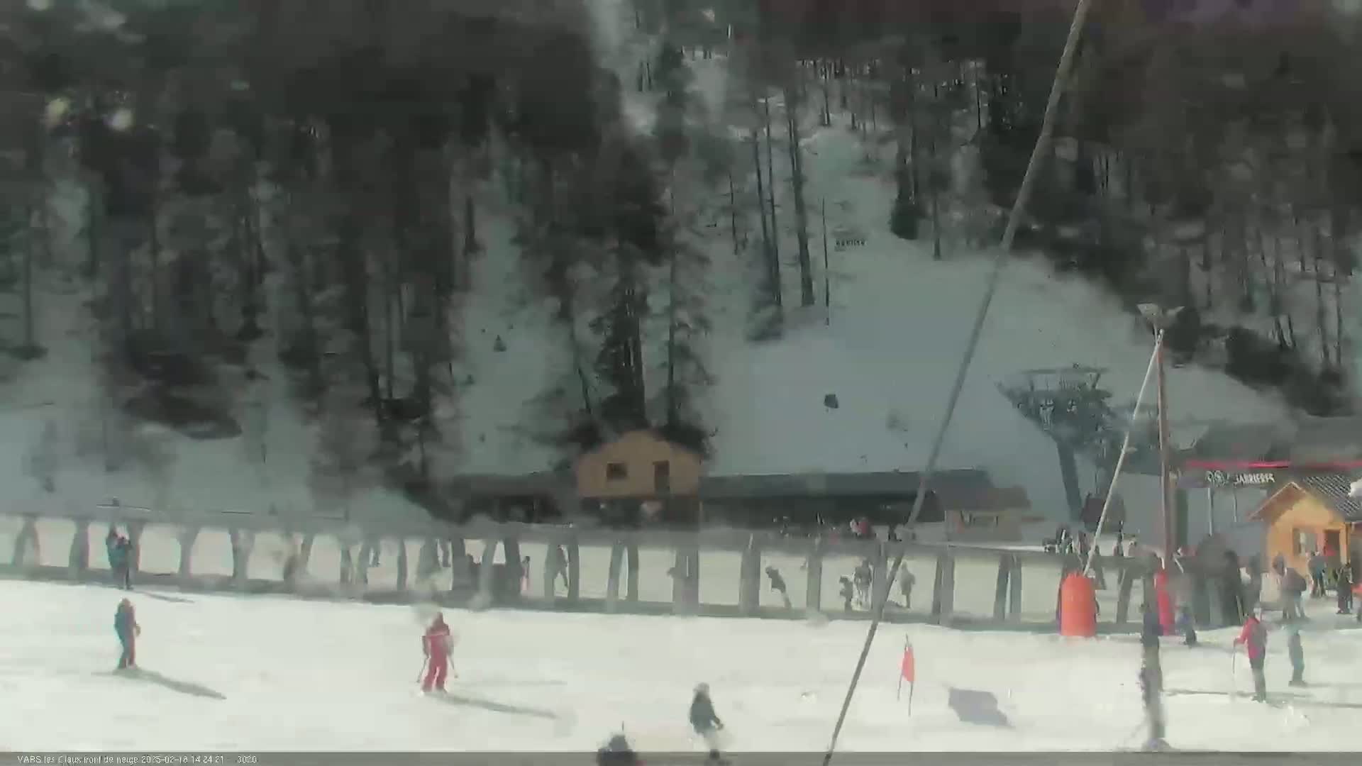 A snowy ski slope with several skiers and snowboarders, a small building, and a wooded area in the background under a bright, sunny sky.