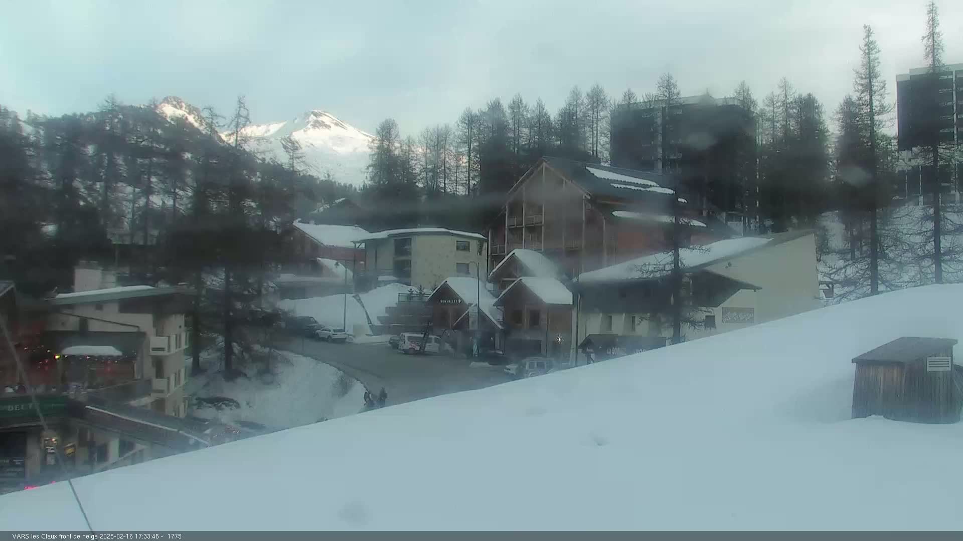 A snowy village scene with several buildings, some cars, and a mountain range in the background under an overcast sky.