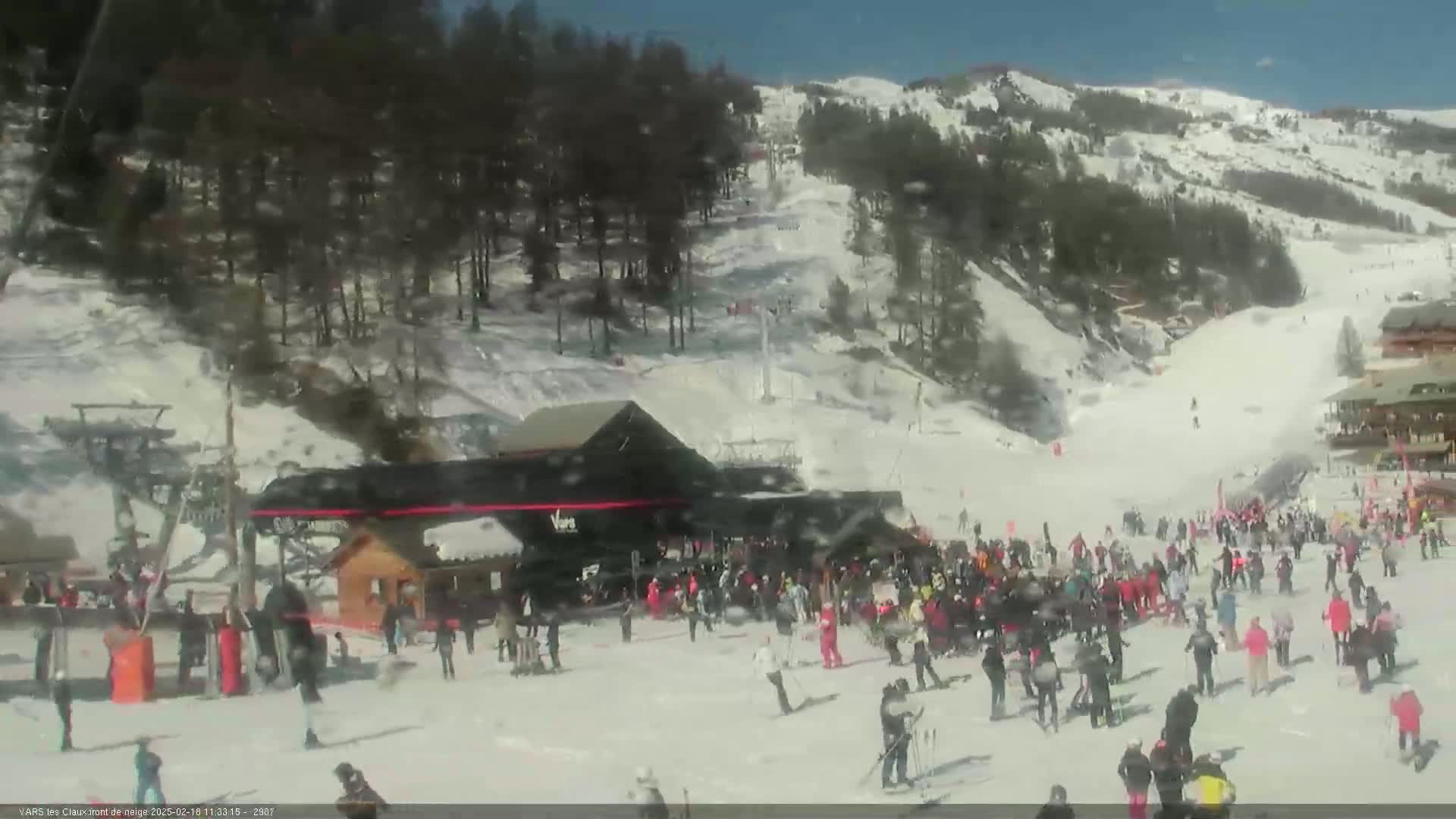 A sunny day at a snow-covered ski resort shows many people gathered near a ski lift base.