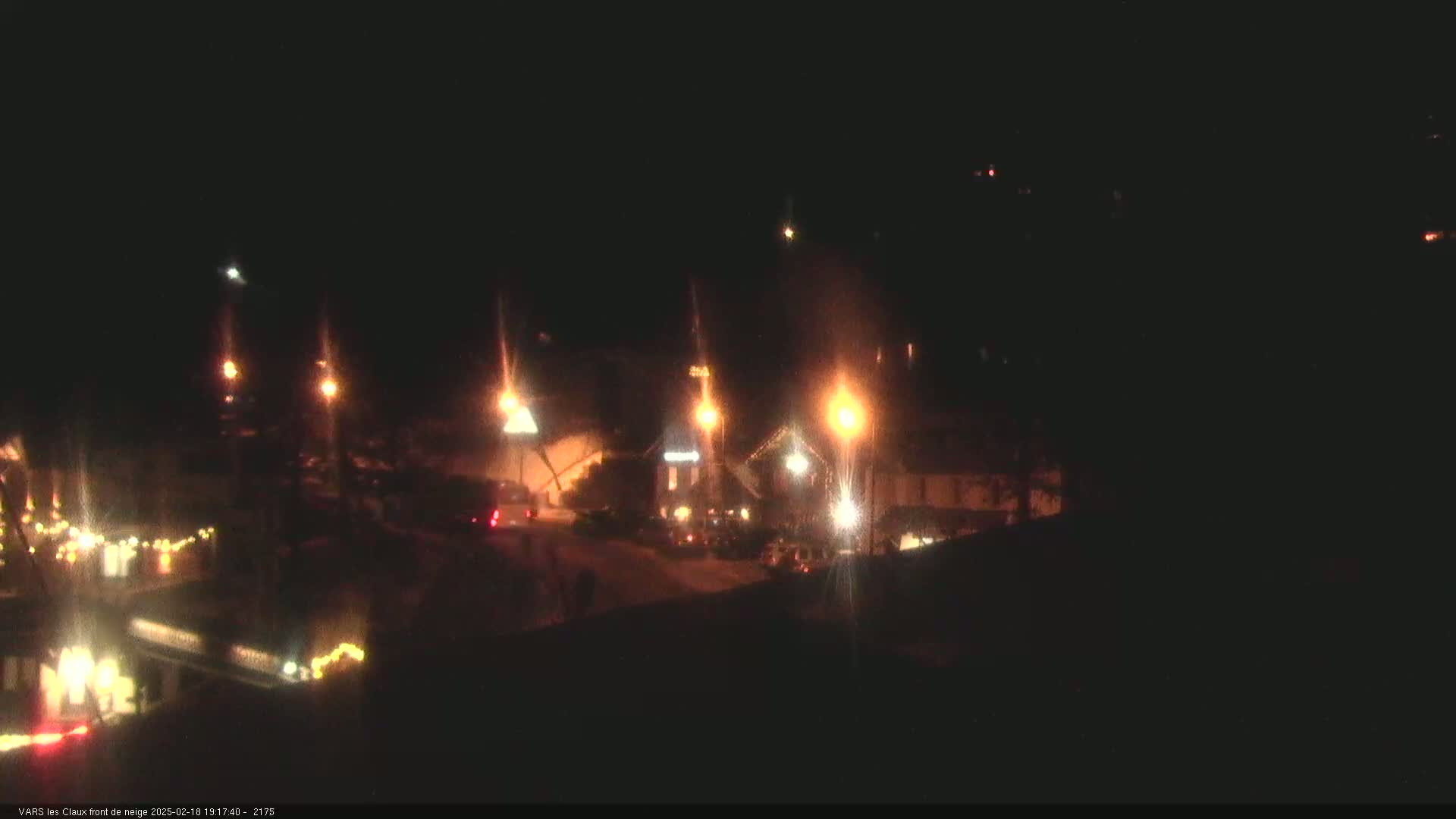 A nighttime street scene shows a few buildings illuminated by streetlights and some vehicles parked along the road, possibly in a small town.