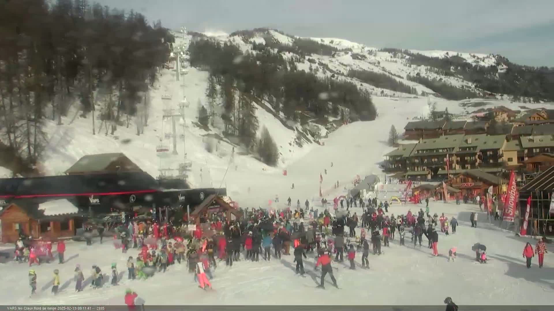 A snowy ski resort with a crowd of people gathered near a building at the base of a ski slope on a partly cloudy day.
