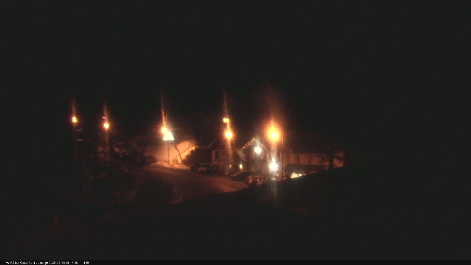 At night, illuminated by several streetlights, a snow-covered road curves past a building complex.
