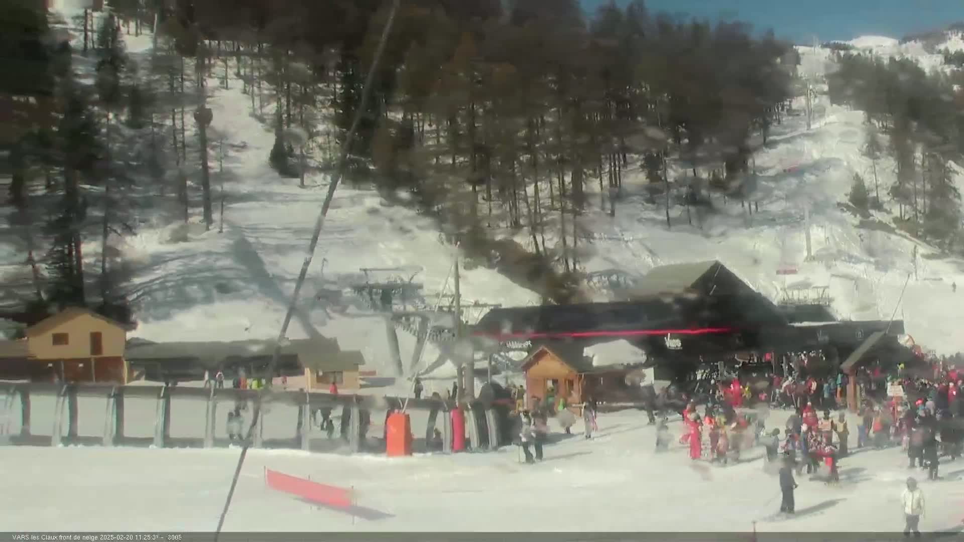 A snowy ski resort on a sunny day shows a crowd of people gathered near the base of a ski lift.