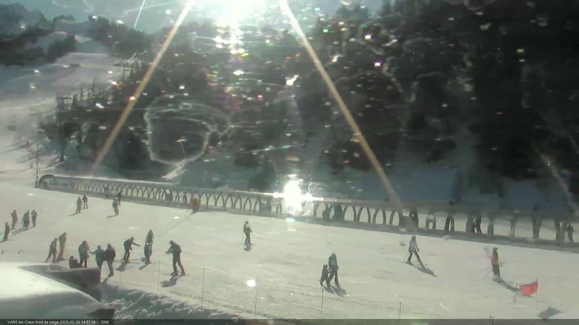 A sunny day on a snow-covered ski slope shows numerous people skiing and standing near a curved fence, with trees in the background.