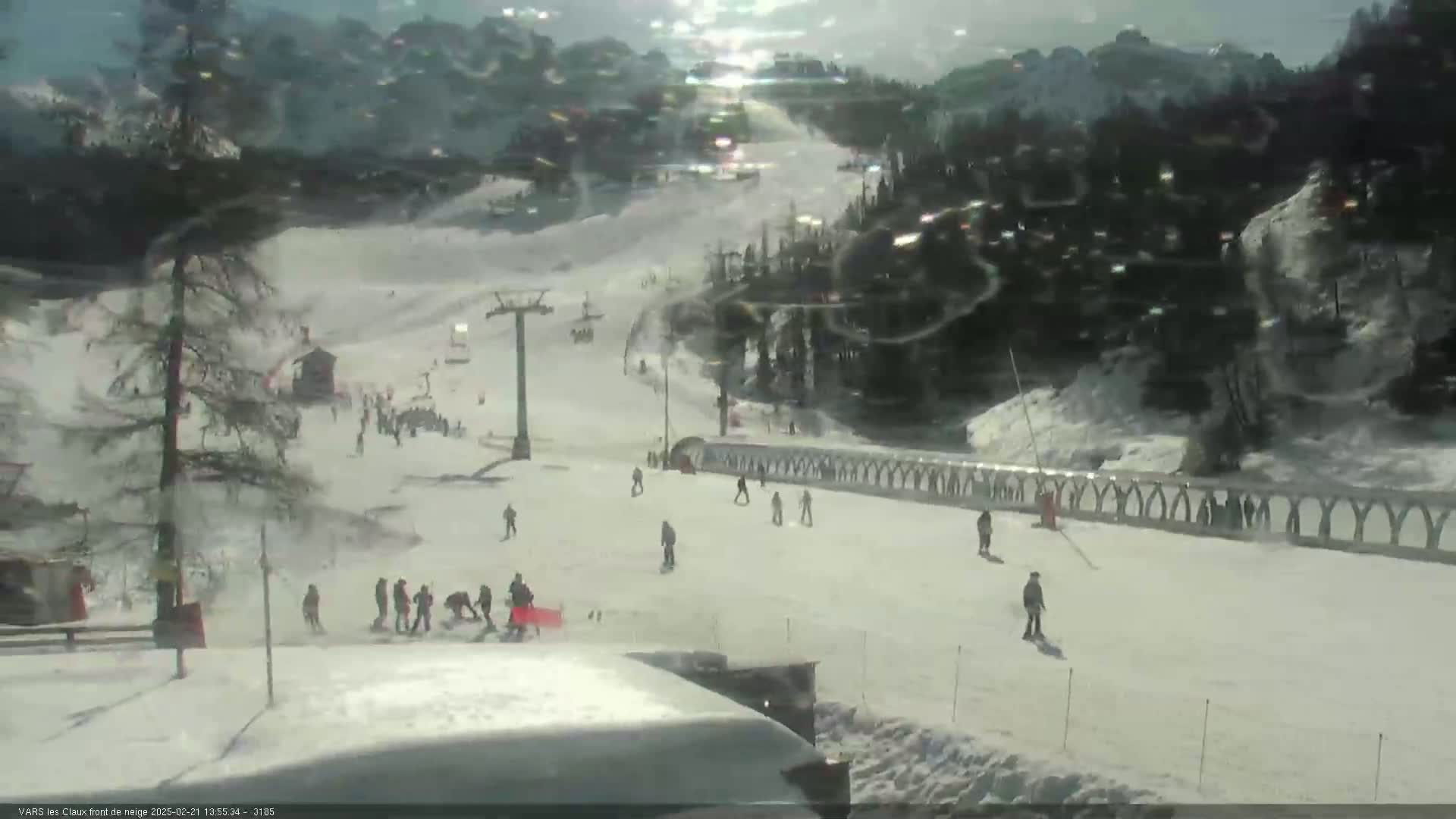 A sunny day at a snow-covered ski resort shows skiers and snowboarders on a slope with a chairlift and snow-covered trees in the background.