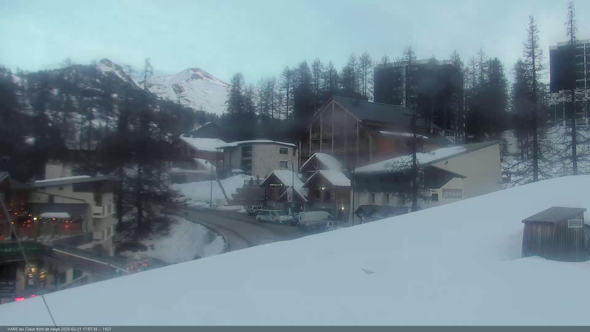A snow-covered mountain village with several buildings, some vehicles on a road, and a mountain in the background under a cloudy sky.