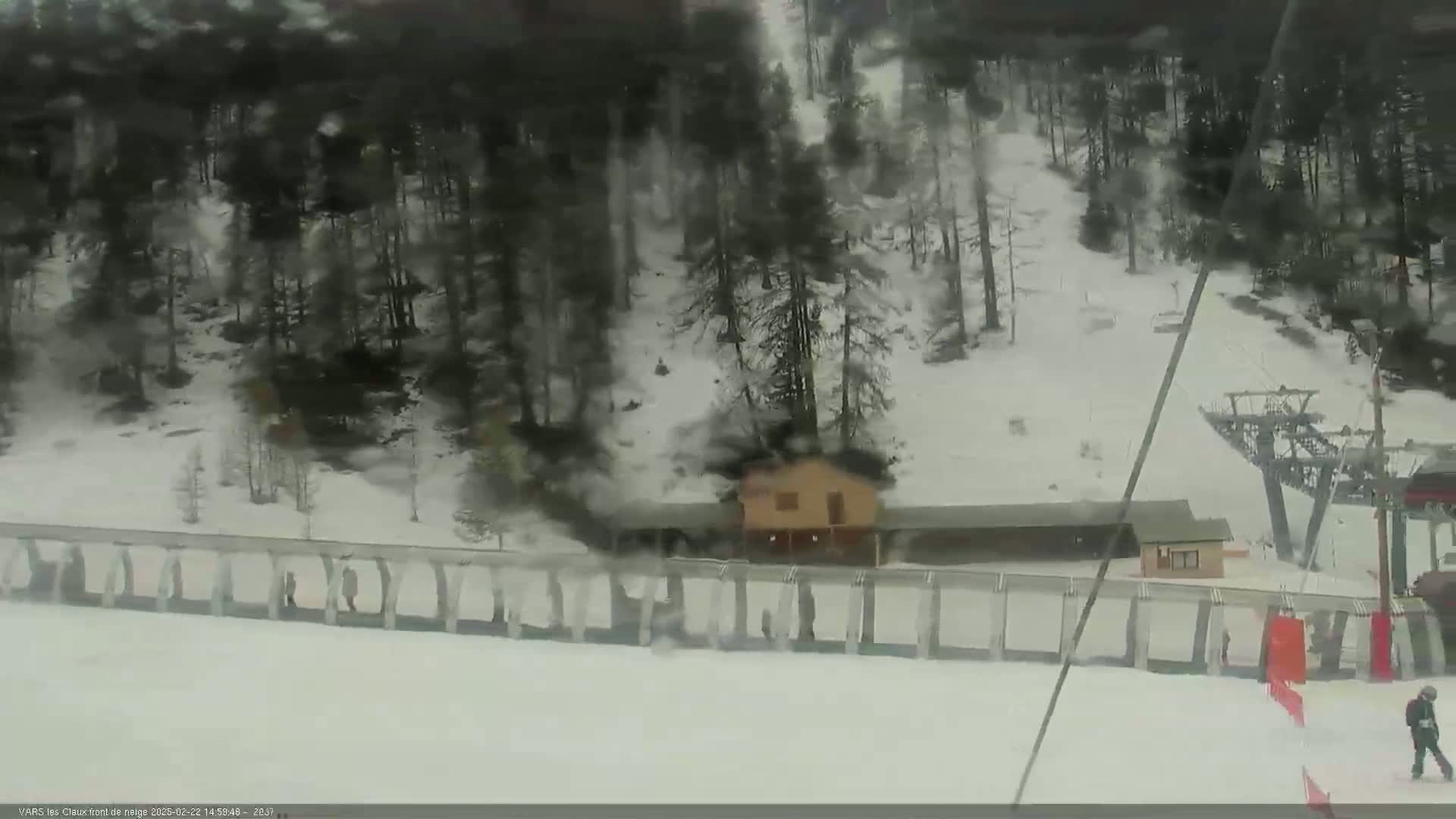A snow-covered ski slope with a small building, a ski lift, and a few people walking near a snow fence.