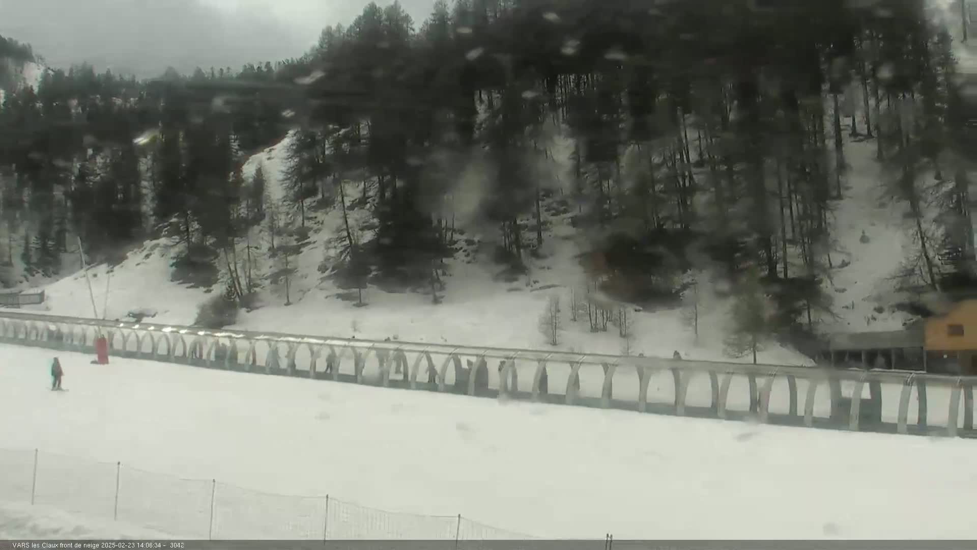 A snowy, overcast scene shows a ski slope with a few people, a long covered walkway, and a dark forest in the background.