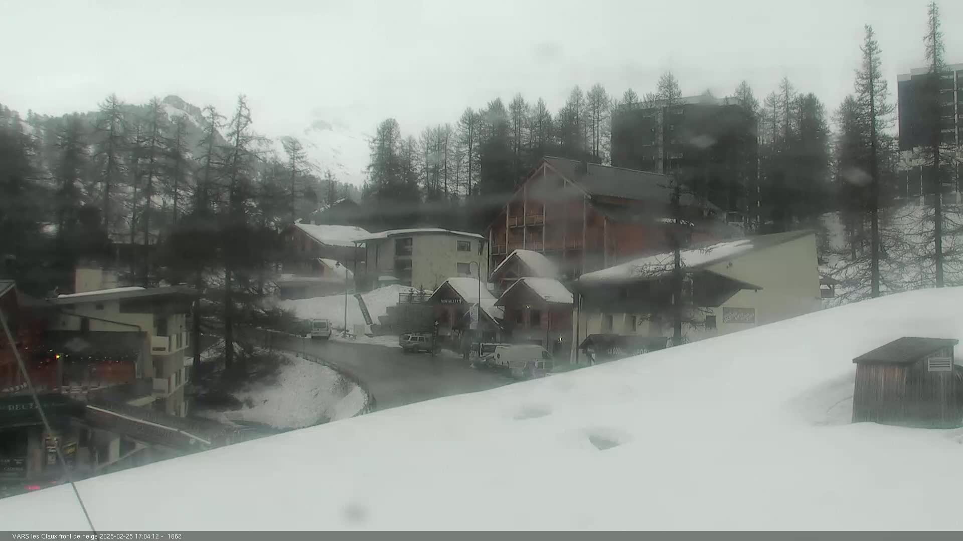 A snow-covered village nestled among evergreen trees on a cloudy day, with a few vehicles visible on a winding road.