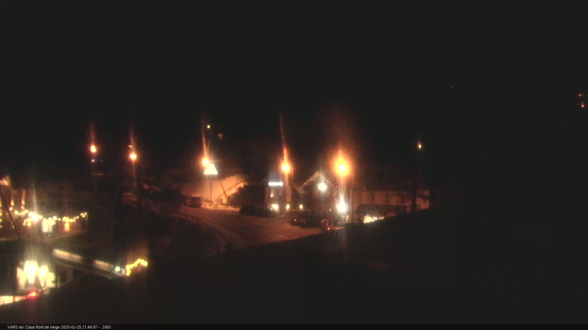 A nighttime view of a snow-covered area with several illuminated buildings and vehicles, streetlights casting a warm glow on the scene.