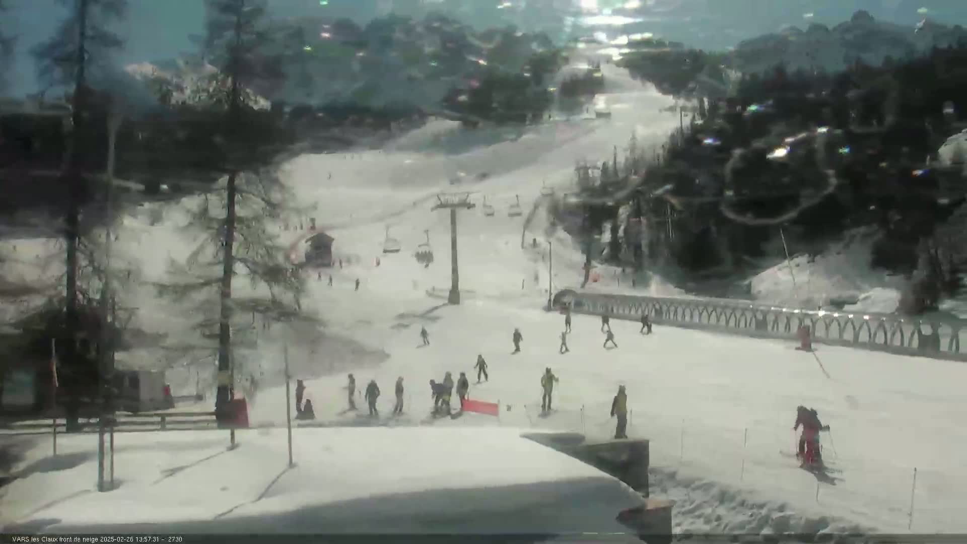 A snowy ski slope with several skiers and snowboarders, a ski lift, and surrounding trees under a bright, sunny sky.