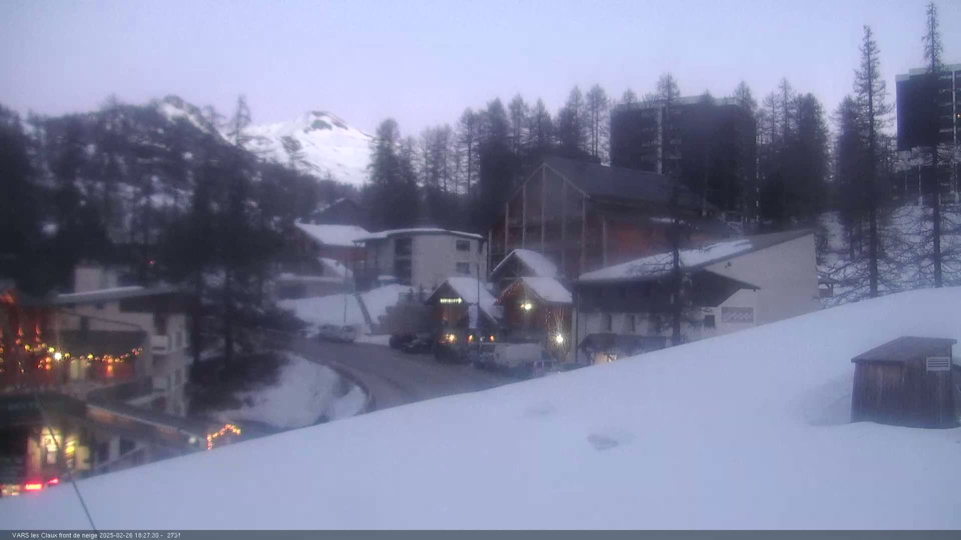 A snow-covered village nestled in the mountains under a twilight sky, with several buildings and vehicles visible.