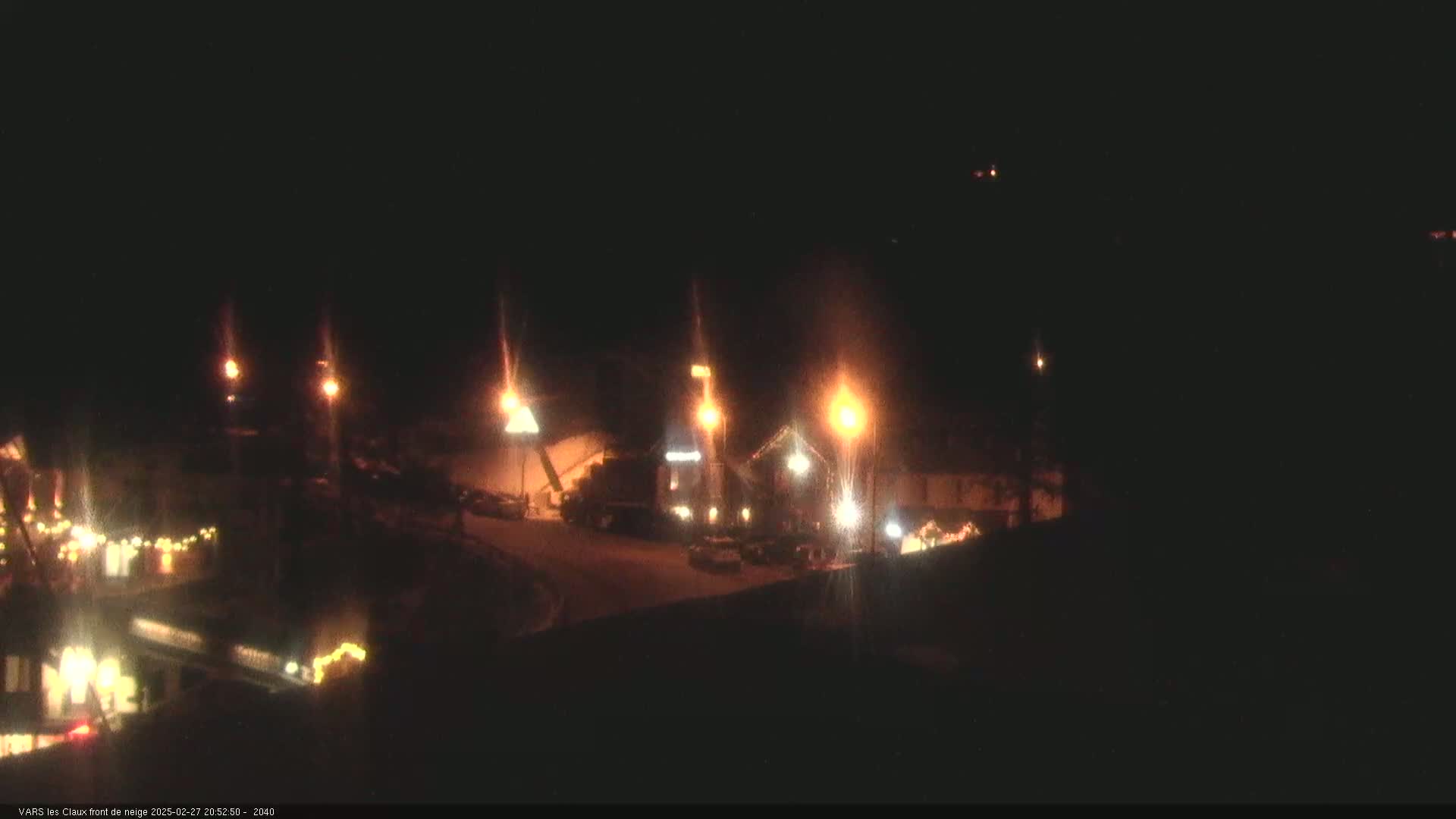 A nighttime, snow-covered scene shows several illuminated buildings and vehicles, with lights casting glare.