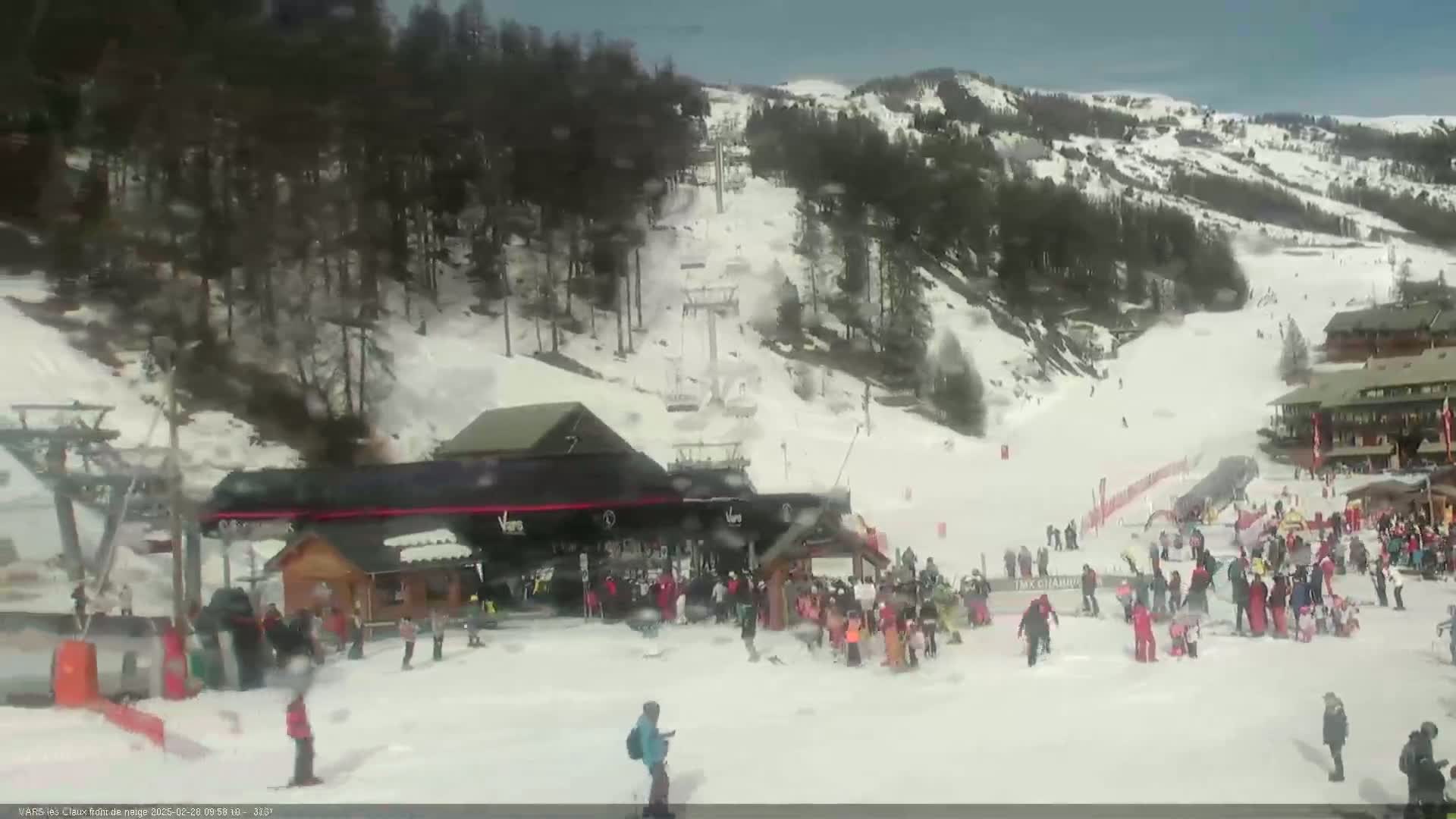 A snowy ski resort with several skiers and snowboarders near a building and chairlifts on a bright, somewhat hazy day.