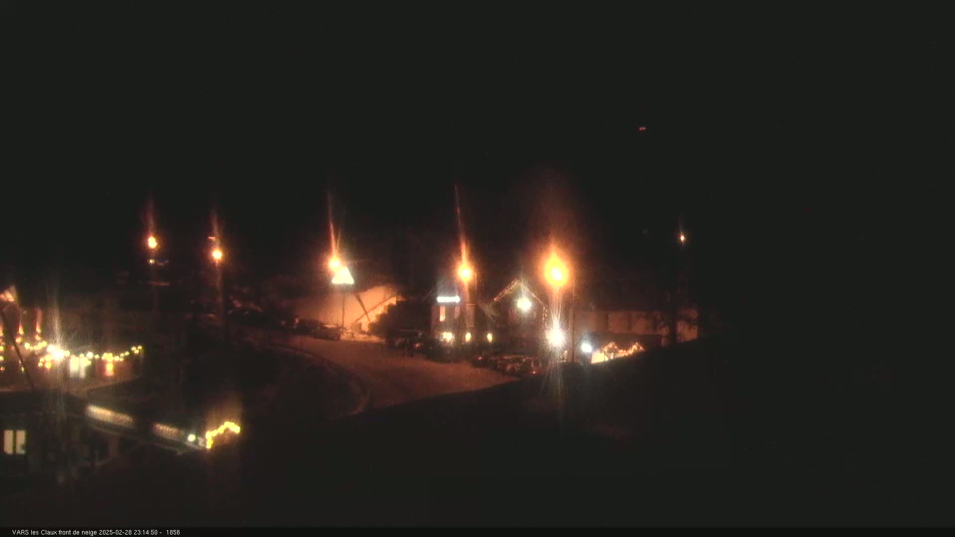 At night, under a dark sky, illuminated buildings and vehicles are visible in a snow-covered area with several streetlights.