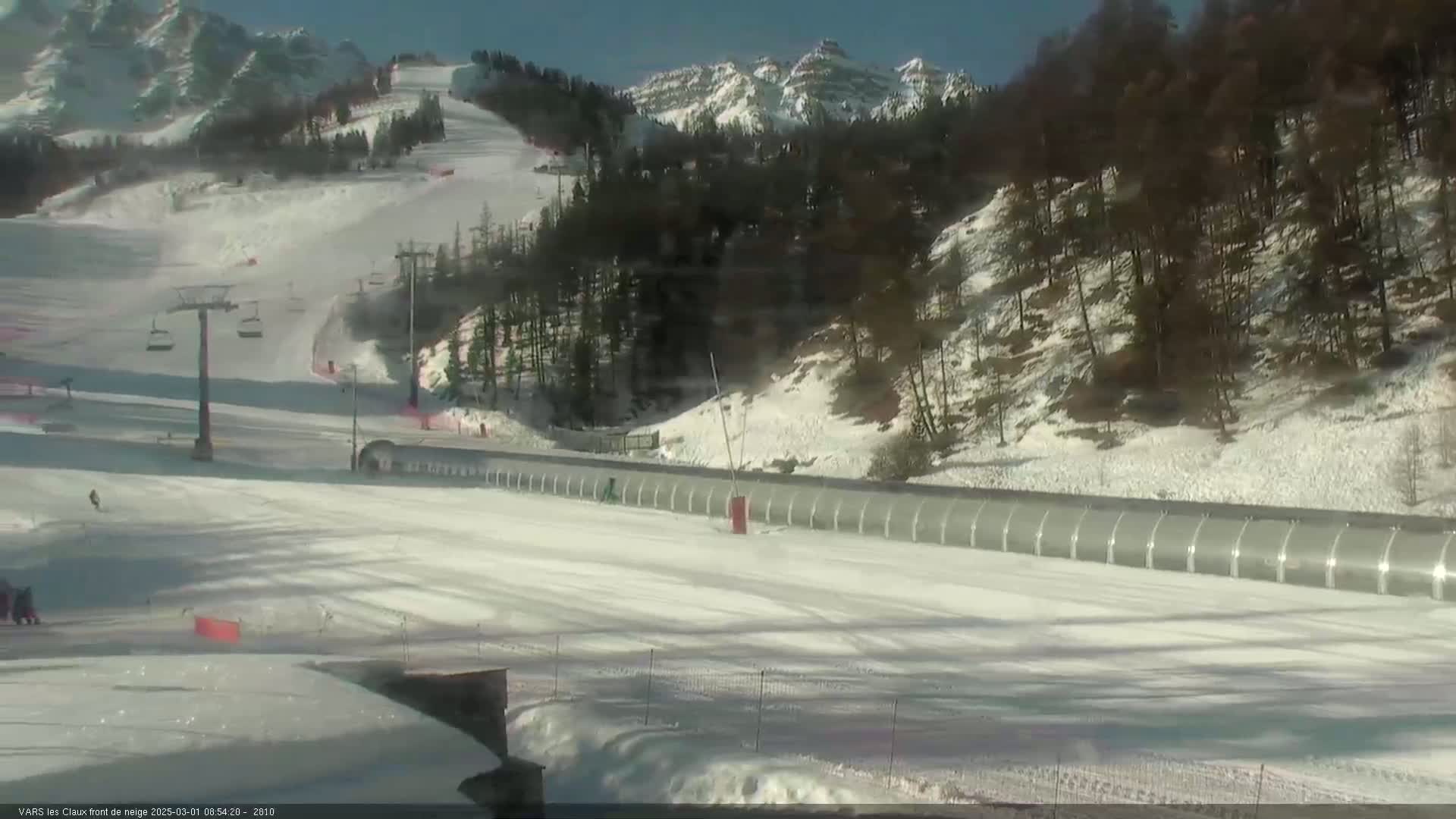 A snow-covered ski slope with a chairlift and a long, covered structure sits beneath sunny skies and snow-dusted mountains.