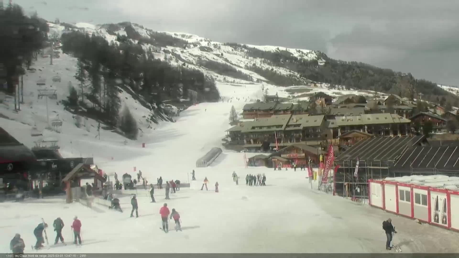 A snow-covered ski slope with several skiers and snowboarders, ski lifts, and buildings under an overcast sky.