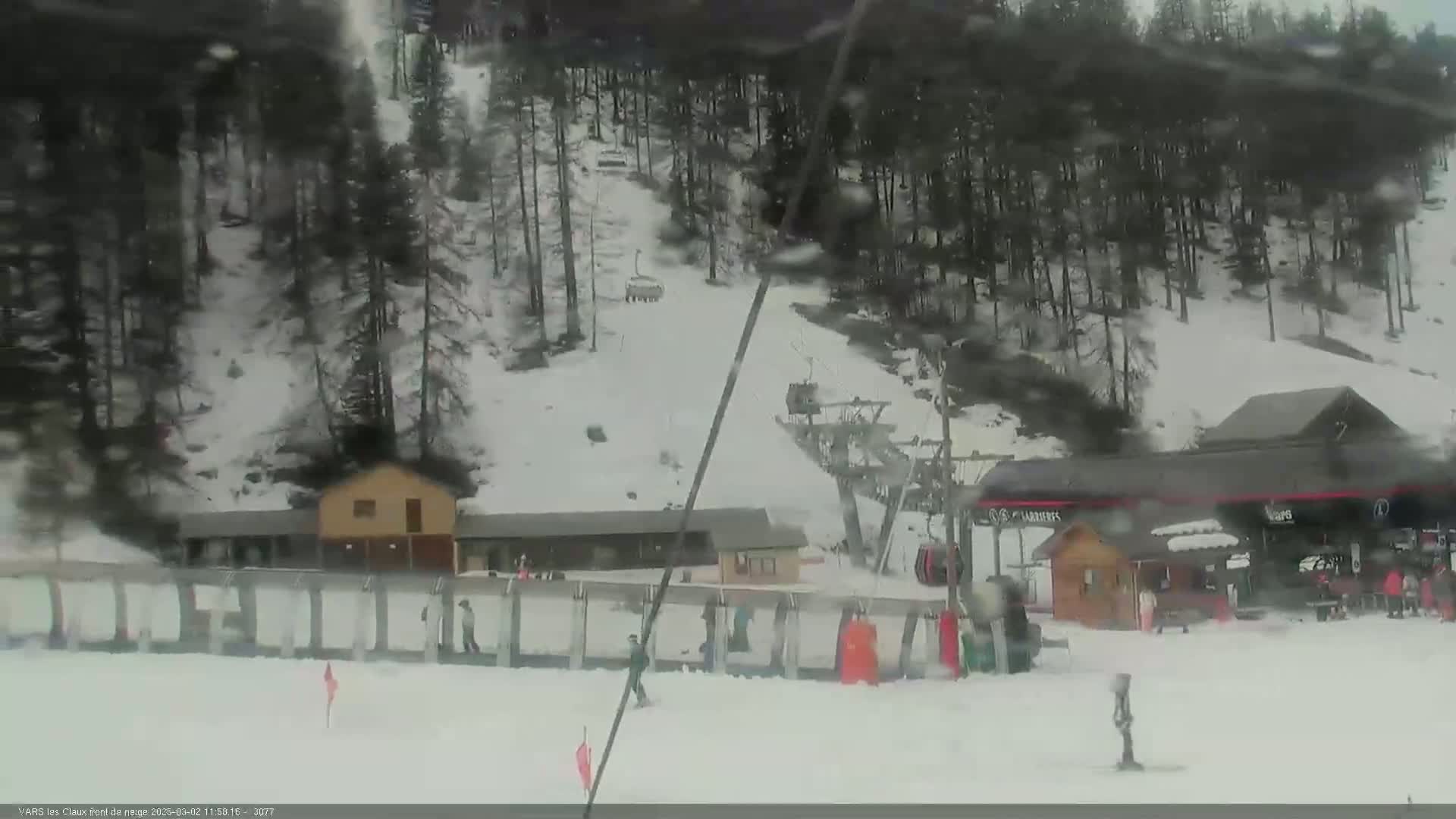 A snowy ski resort scene shows a chairlift, several small buildings, and skiers on a slope under overcast skies.
