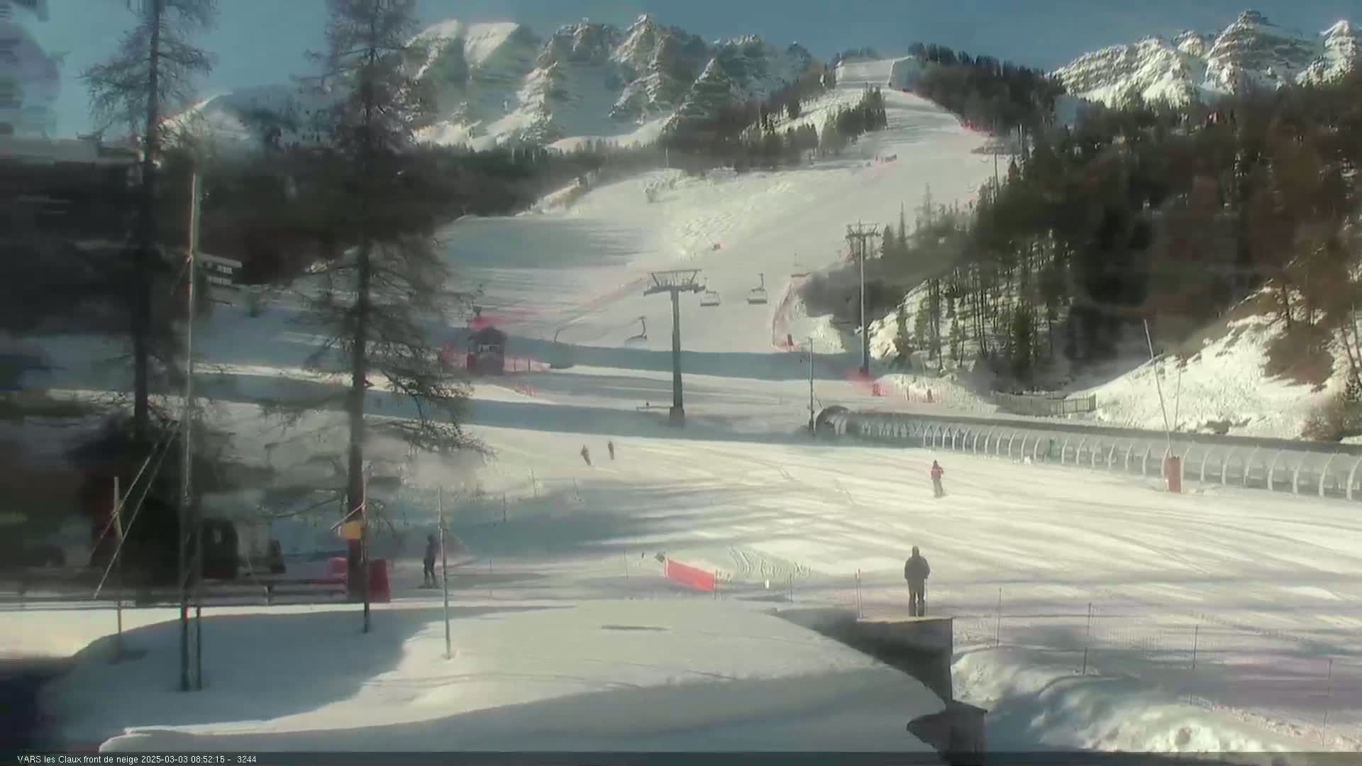 A sunny, snow-covered ski slope with a chairlift and several skiers visible against a backdrop of snow-capped mountains.