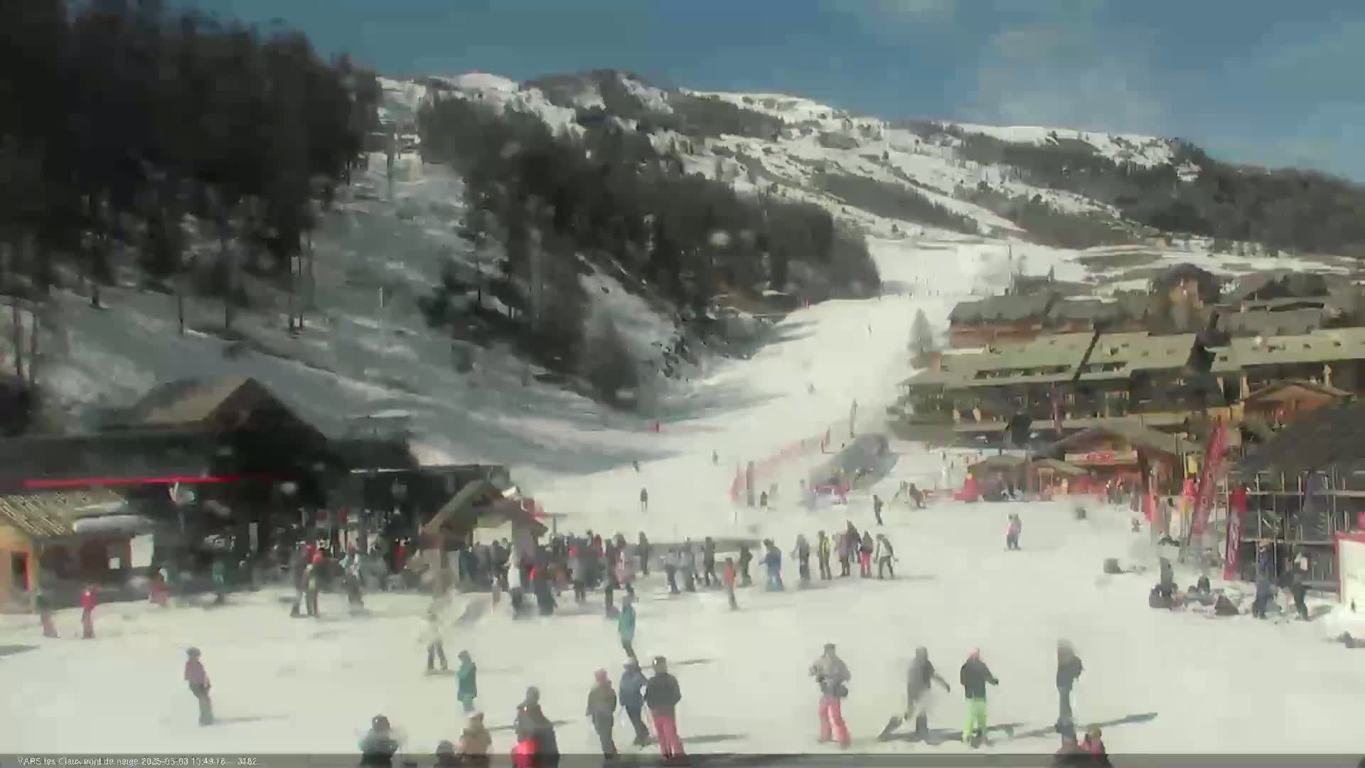 A sunny day at a snow-covered ski resort shows skiers and snowboarders at the bottom of a slope near several buildings.