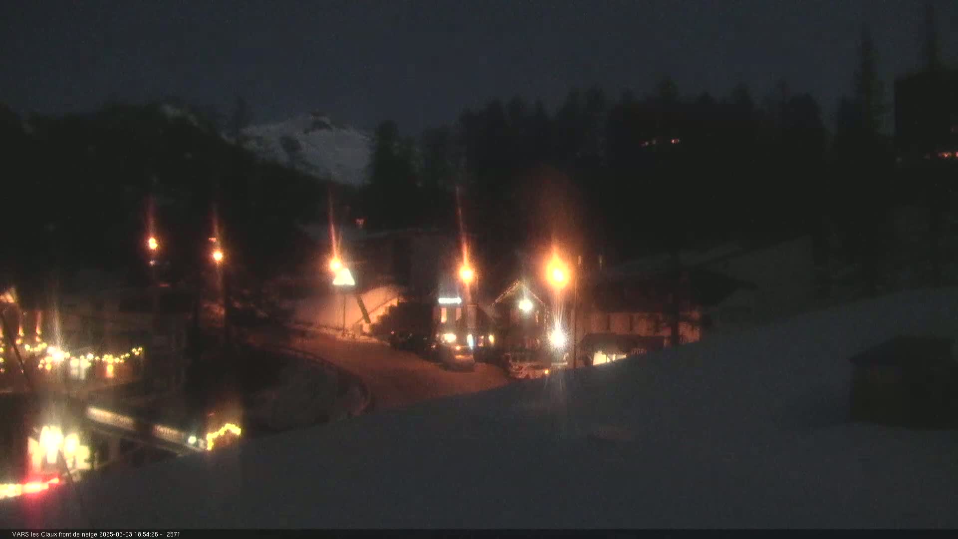 A snow-covered landscape at night, illuminated by streetlights, shows a few buildings and vehicles.
