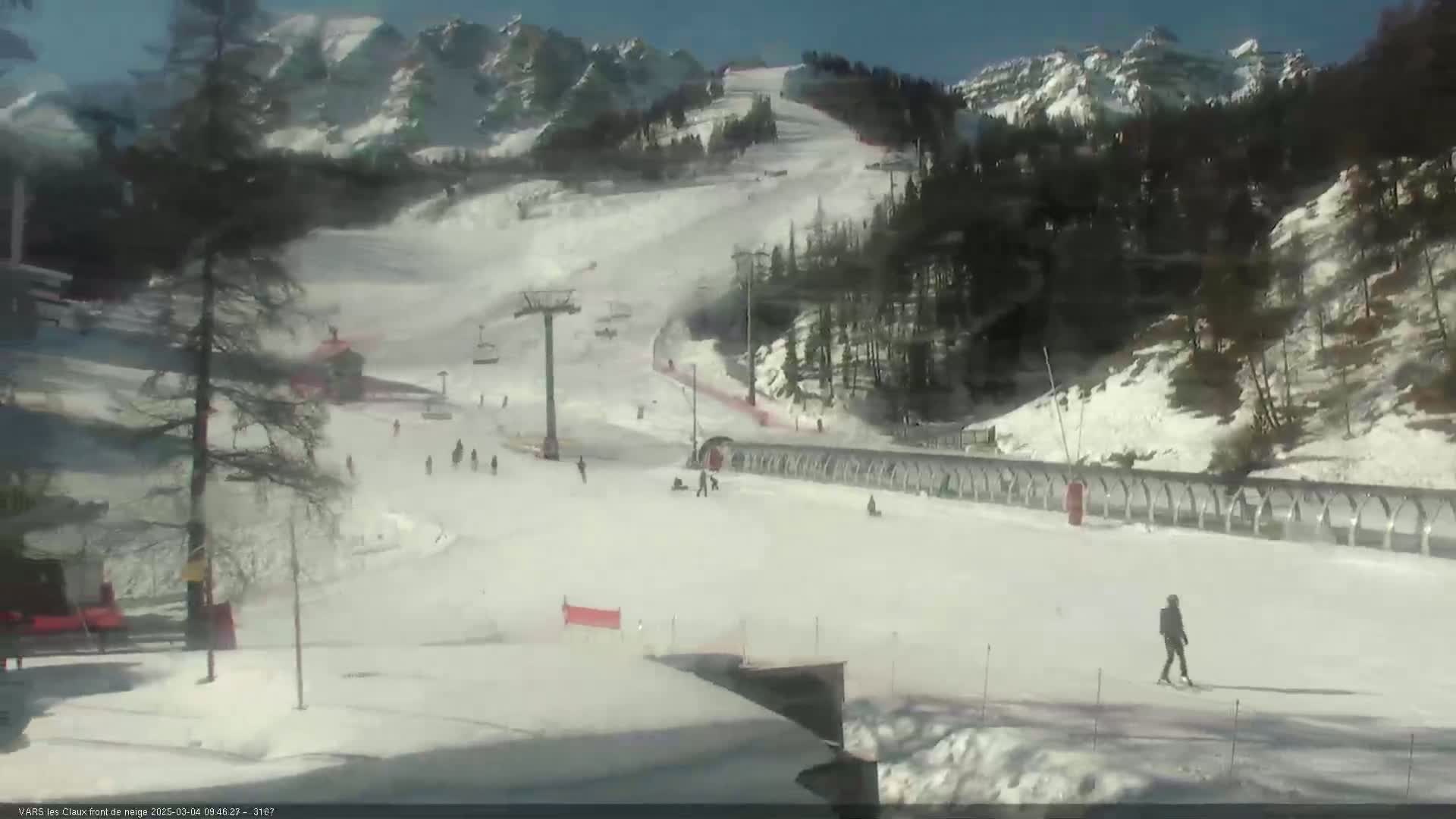 A sunny, snow-covered ski slope with a chairlift, skiers, and snowboarders, is set against a backdrop of snow-capped mountains and evergreen trees.