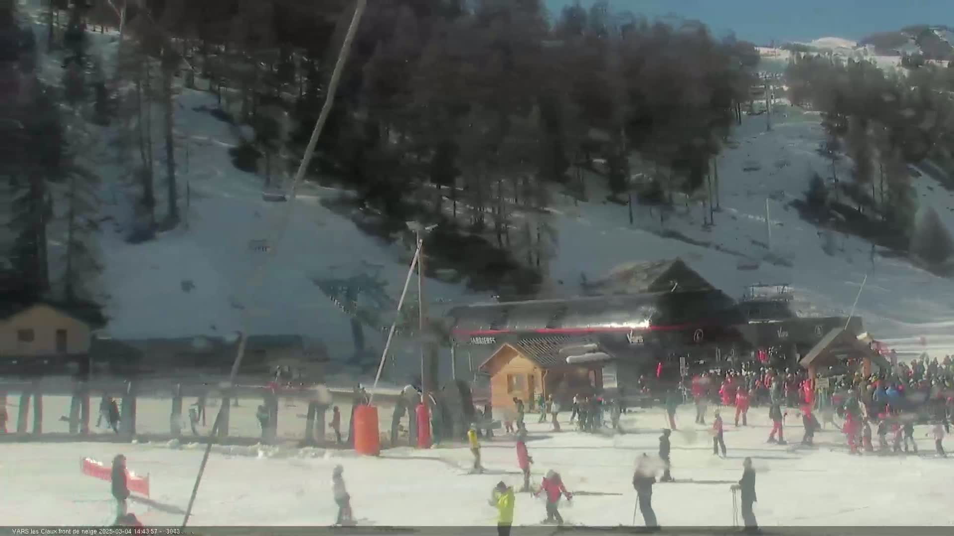 A sunny, snow-covered ski slope with skiers and snowboarders near a ski lift and lodge.
