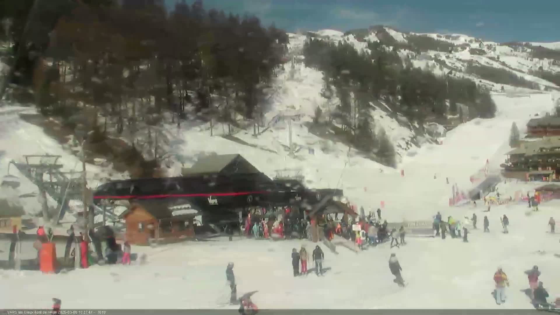A ski resort on a sunny day shows skiers and snowboarders on snowy slopes, with a lift station and buildings at the bottom.
