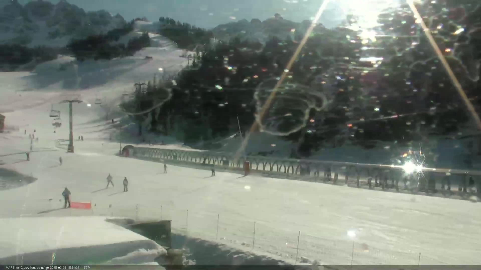 A sunny day at a snow-covered ski resort shows several skiers on a slope with a ski lift and a snow-covered mountain in the background.