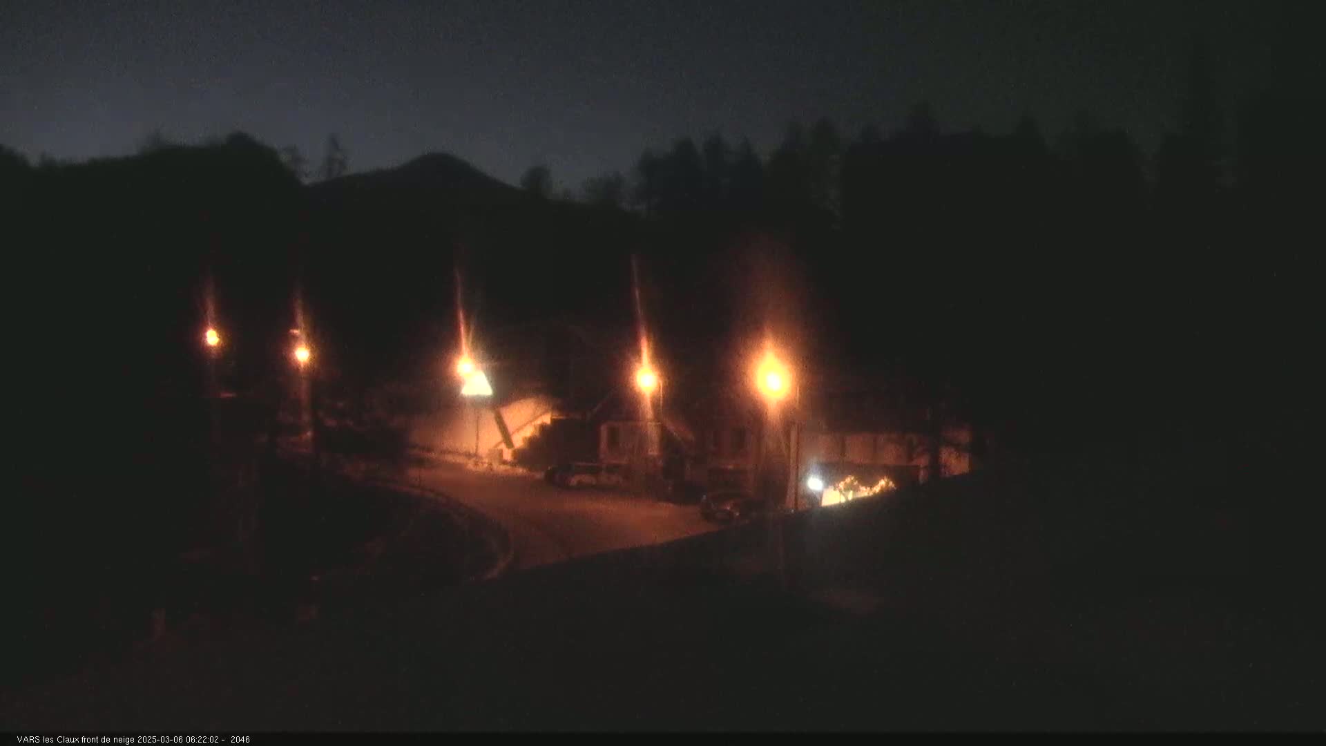 At night, several illuminated structures are visible near a dark, tree-lined hill.