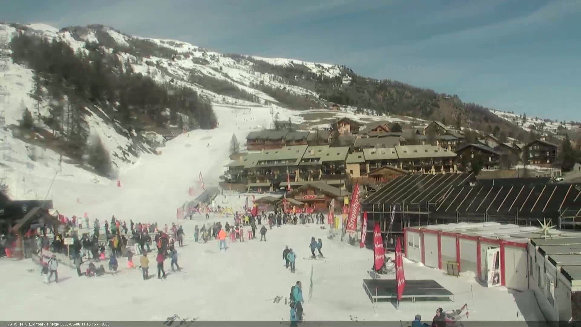 A snow-covered ski resort base area with many people, buildings, and a ski slope on a sunny day.