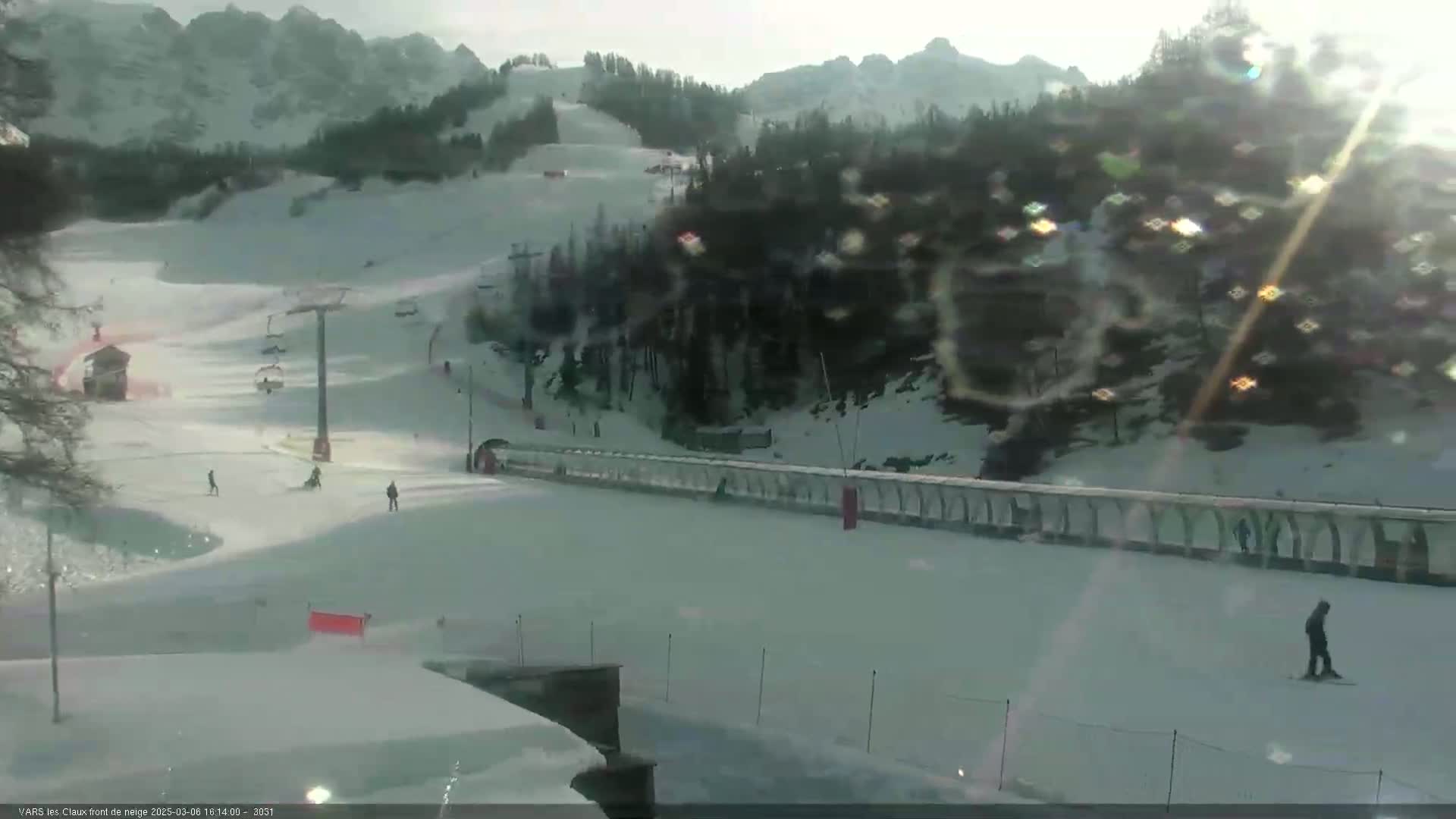 A snowy ski slope with a chairlift and several skiers visible under a bright, sunny sky.
