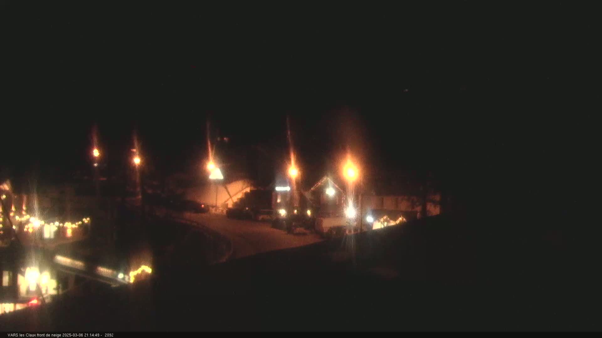 At night, under a clear dark sky, illuminated buildings and streetlights are visible in a snow-covered area with decorative lights.