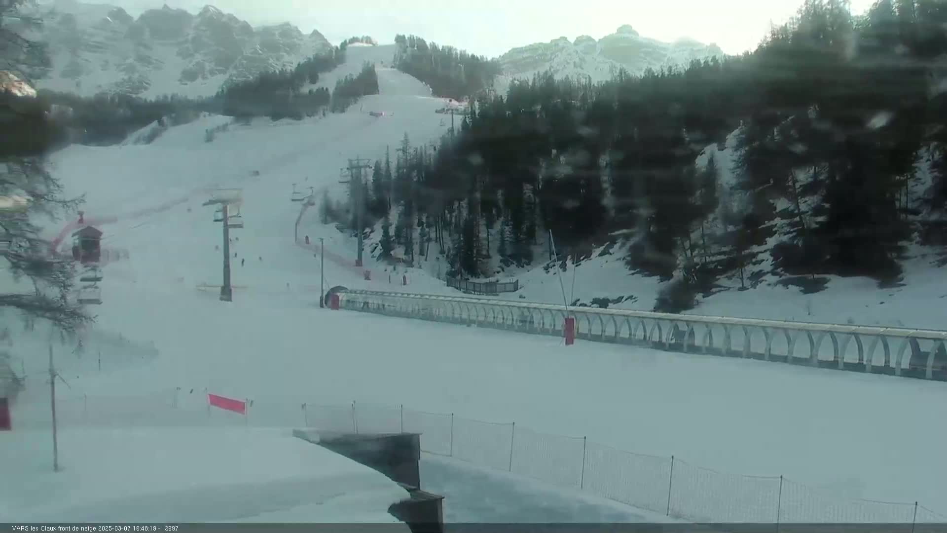 A snow-covered ski slope with several ski lifts and a long, covered conveyor belt is visible on a somewhat overcast day in a mountainous region.