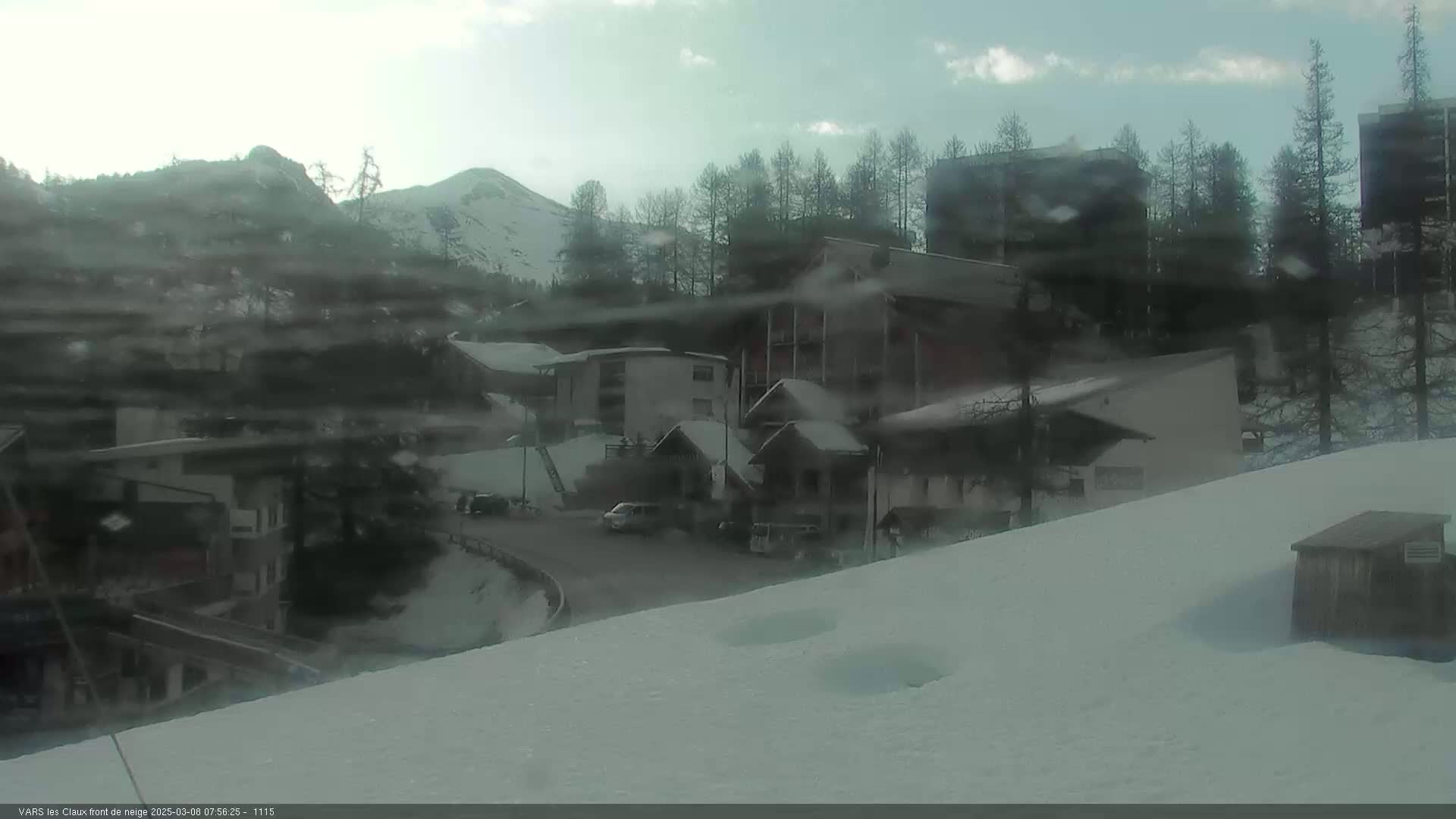 A snowy mountain village scene with overcast skies shows several buildings, a few cars, and some evergreens.