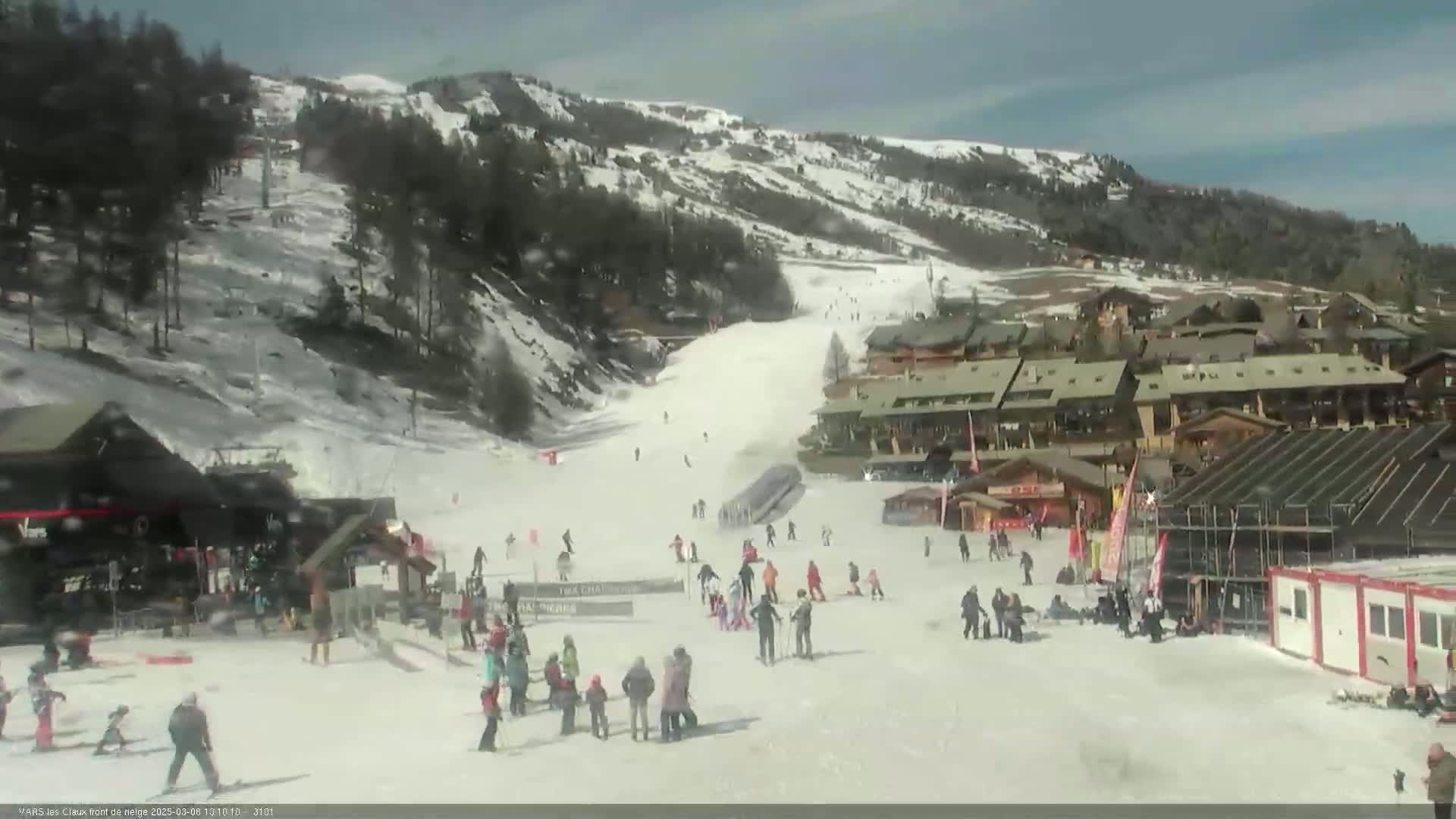A snowy ski slope with skiers and snowboarders, a cluster of buildings, and a partly sunny sky.