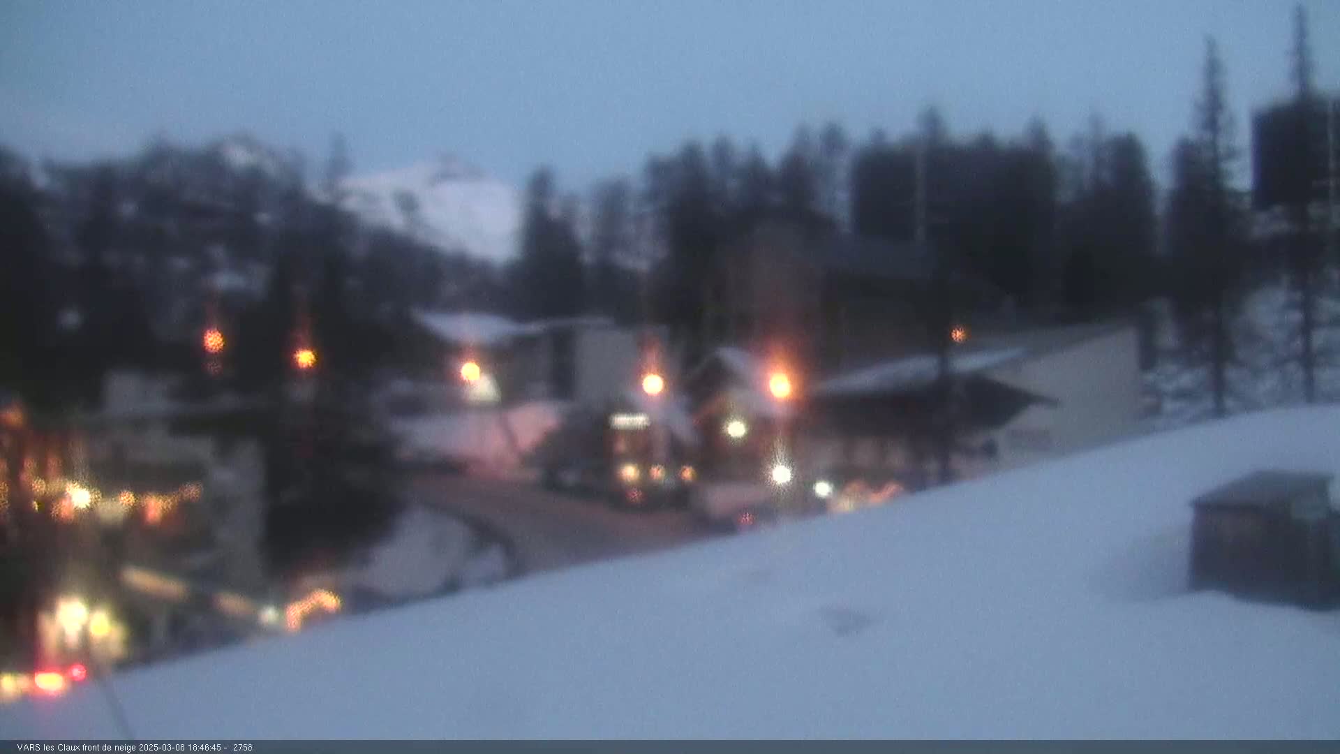 A blurry nighttime view of a snow-covered area with buildings and vehicles, illuminated by scattered lights under a twilight sky.