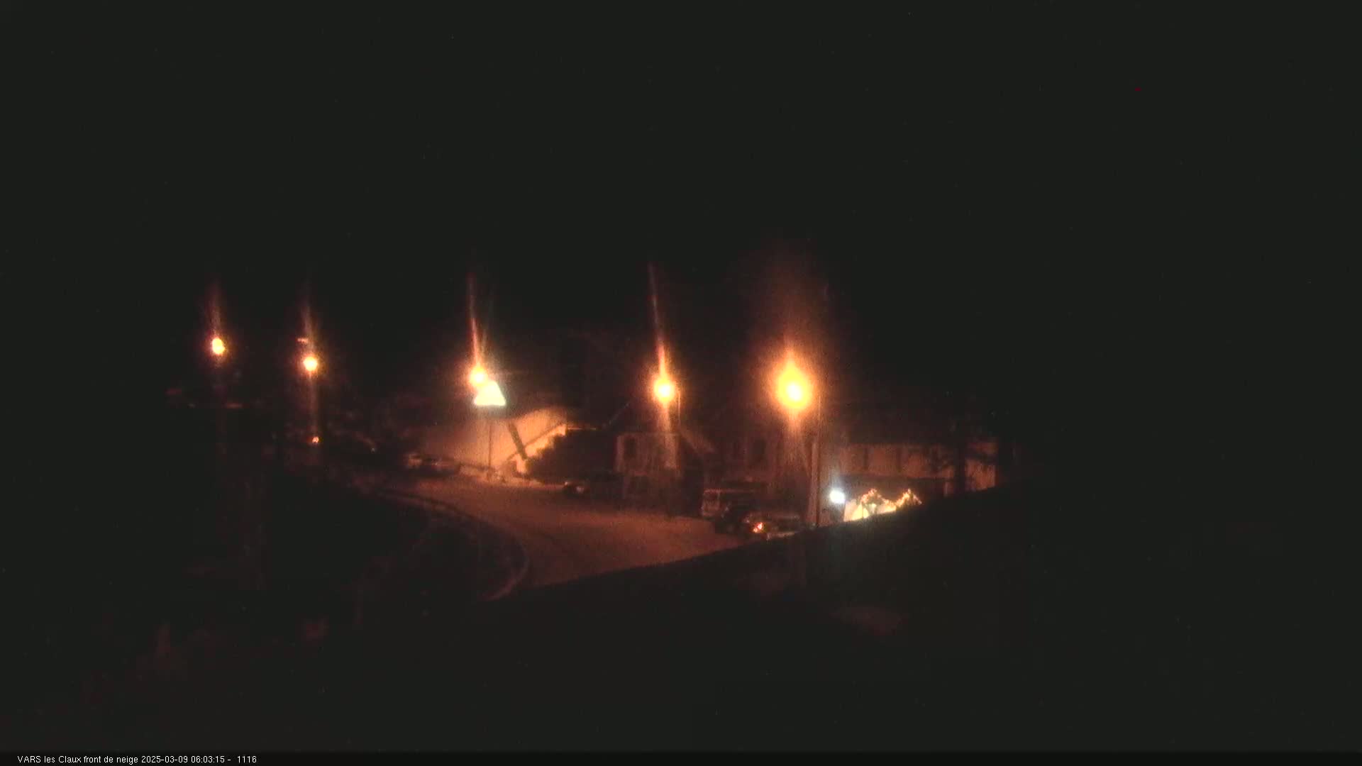 At night, several dimly lit streetlights illuminate a dark, snow-covered area with some vehicles and buildings.