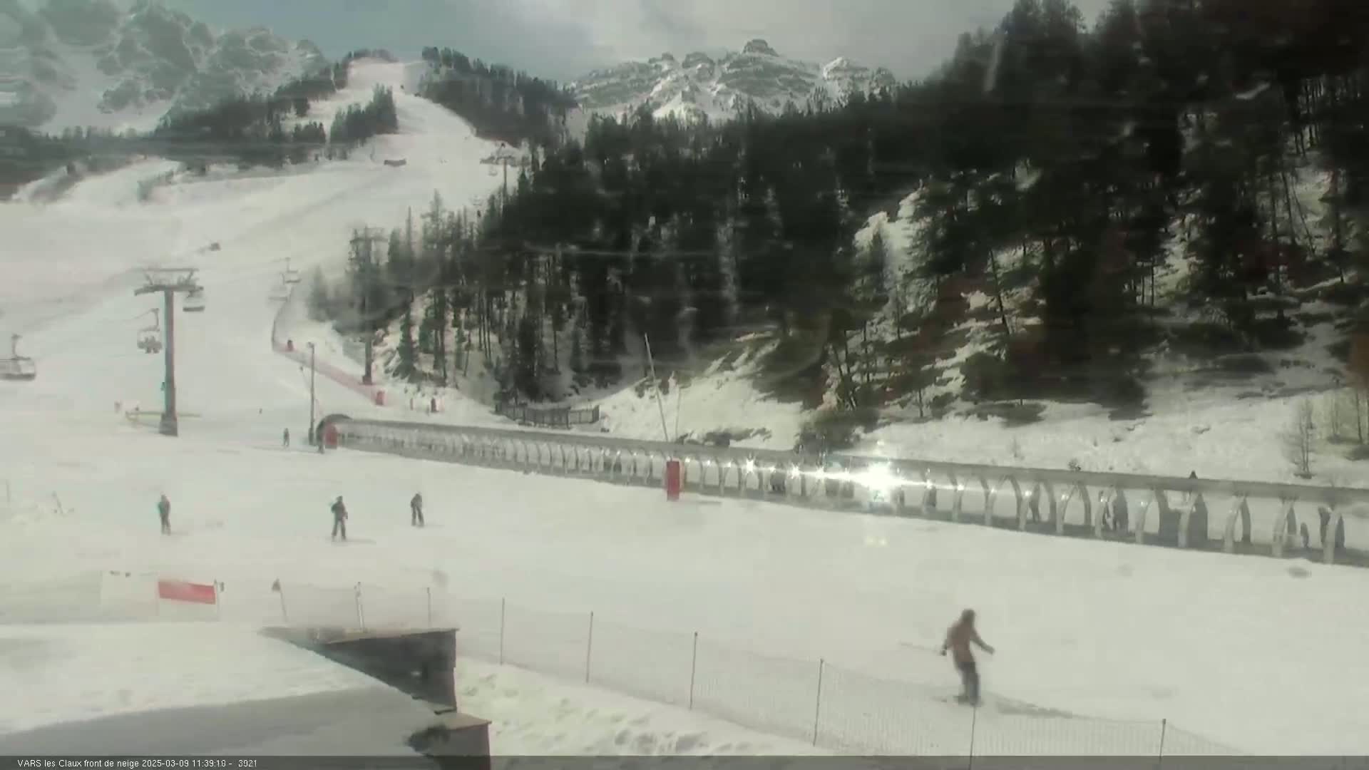 A snowy ski slope with several skiers and a ski lift, under an overcast sky, is situated against a backdrop of snow-covered mountains and evergreen trees.